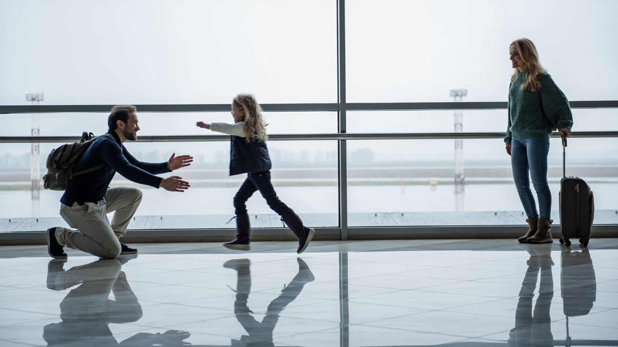 A father kneels in an airport terminal, arms open wide, as his daughter runs toward him. The mother stands nearby, holding a suitcase, smiling at their joyful reunion.