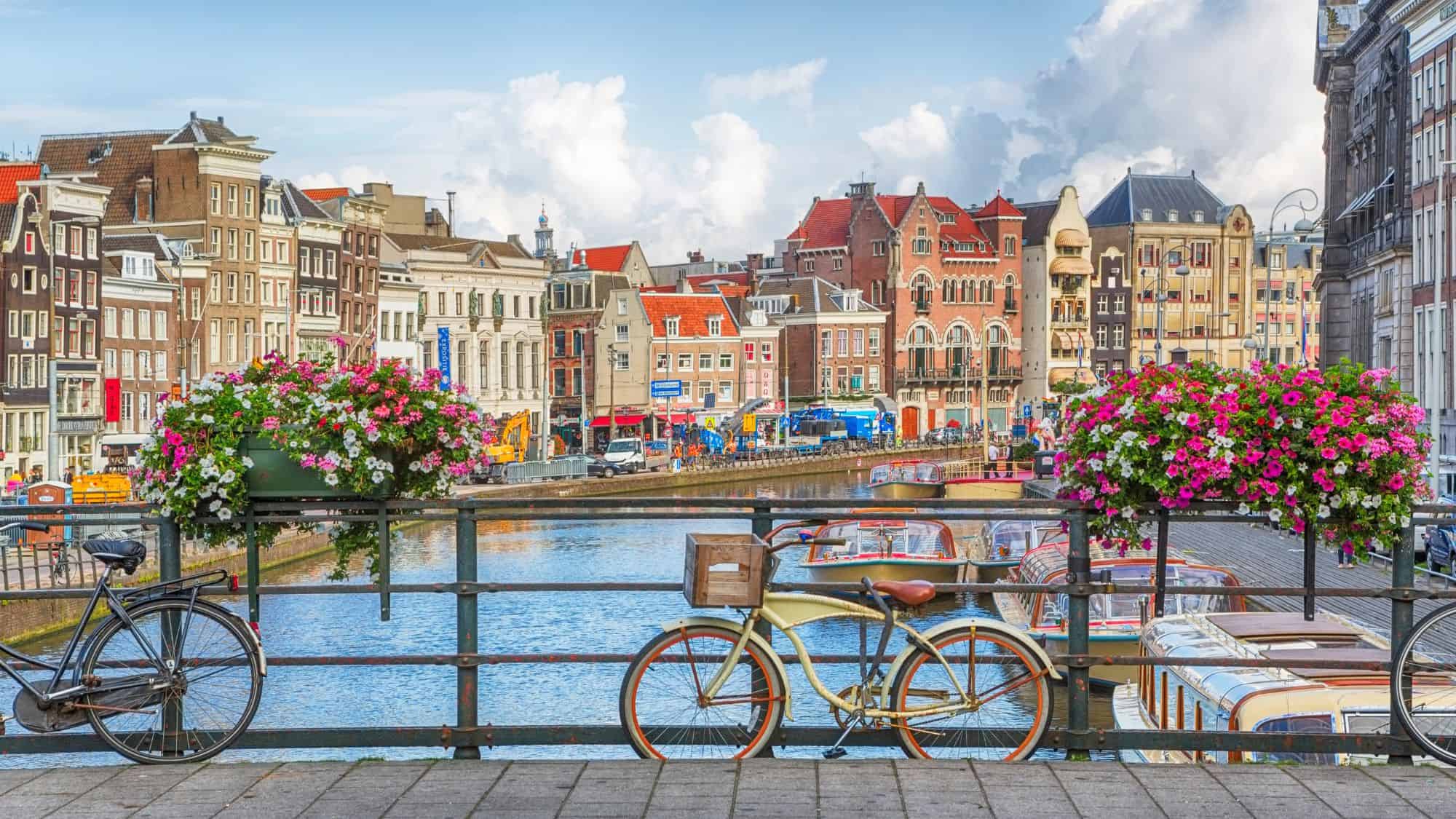 A vibrant view of an Amsterdam canal with colorful, historic buildings lining the waterway. Two bicycles lean against a railing adorned with blooming pink and white flowers, while canal boats float in the background under a partly cloudy sky.
