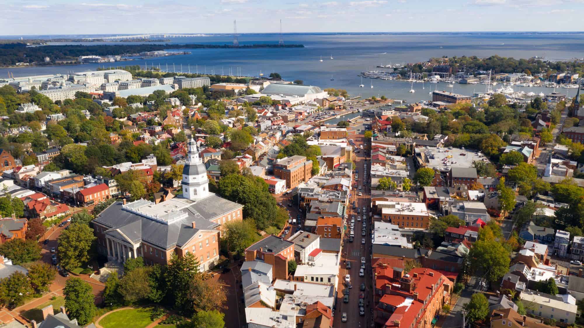 An aerial view of Annapolis, Maryland, showcasing historic architecture, tree-lined streets, and the iconic Maryland State House. The Chesapeake Bay and a marina filled with sailboats stretch out in the background.