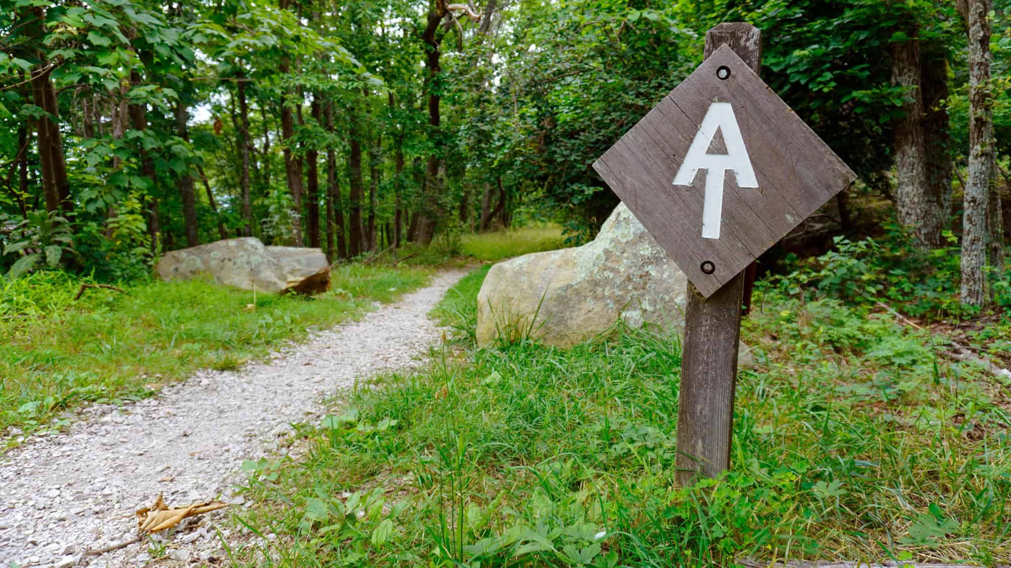 A wooden signpost along a gravel hiking path in a lush green forest, marked with the iconic "AT" symbol, indicating the Appalachian Trail.
