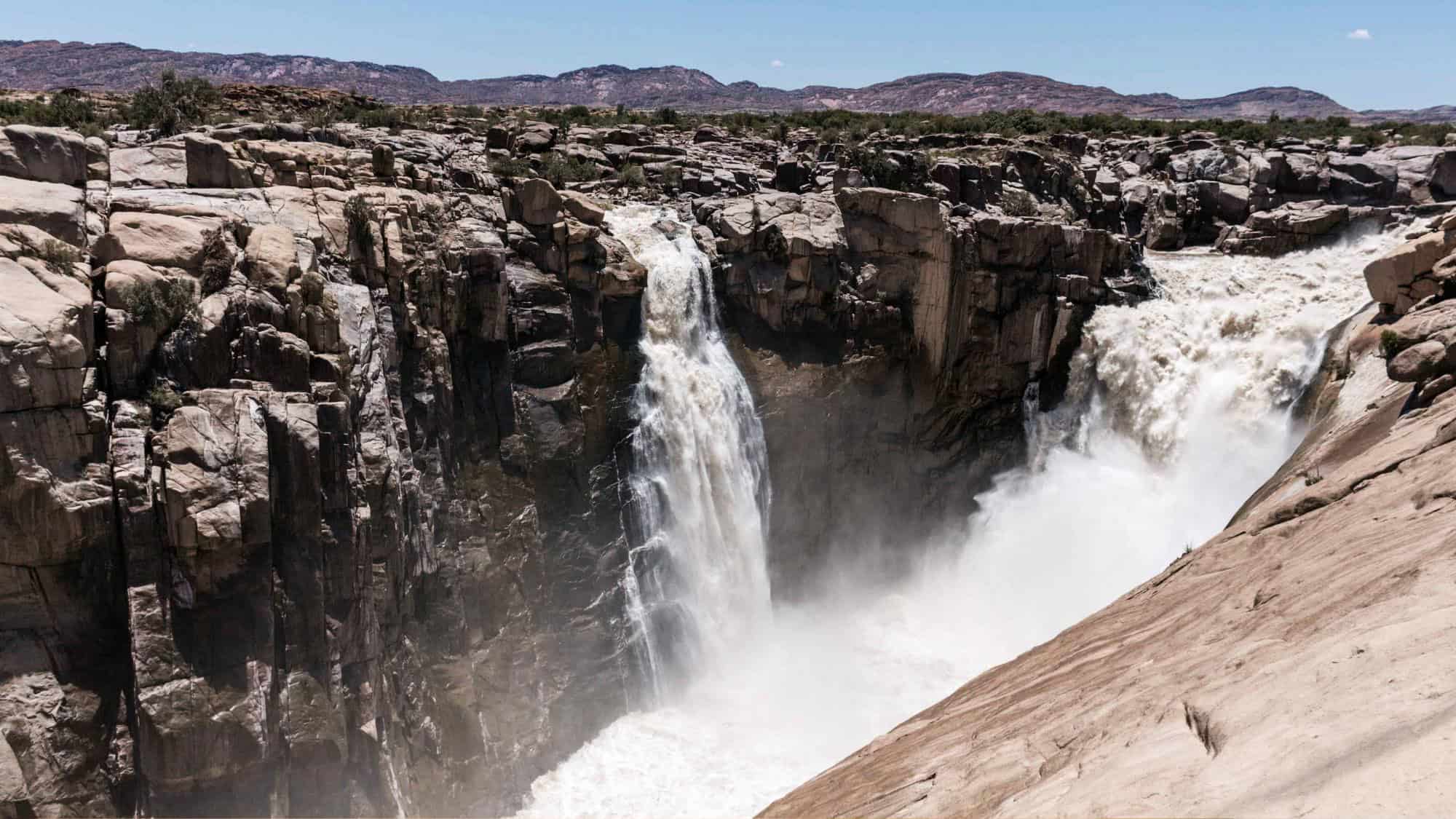 A powerful waterfall plunges down a rocky cliffside, sending up a misty spray. The surrounding rugged terrain and rolling hills in the background highlight the area's raw natural beauty.