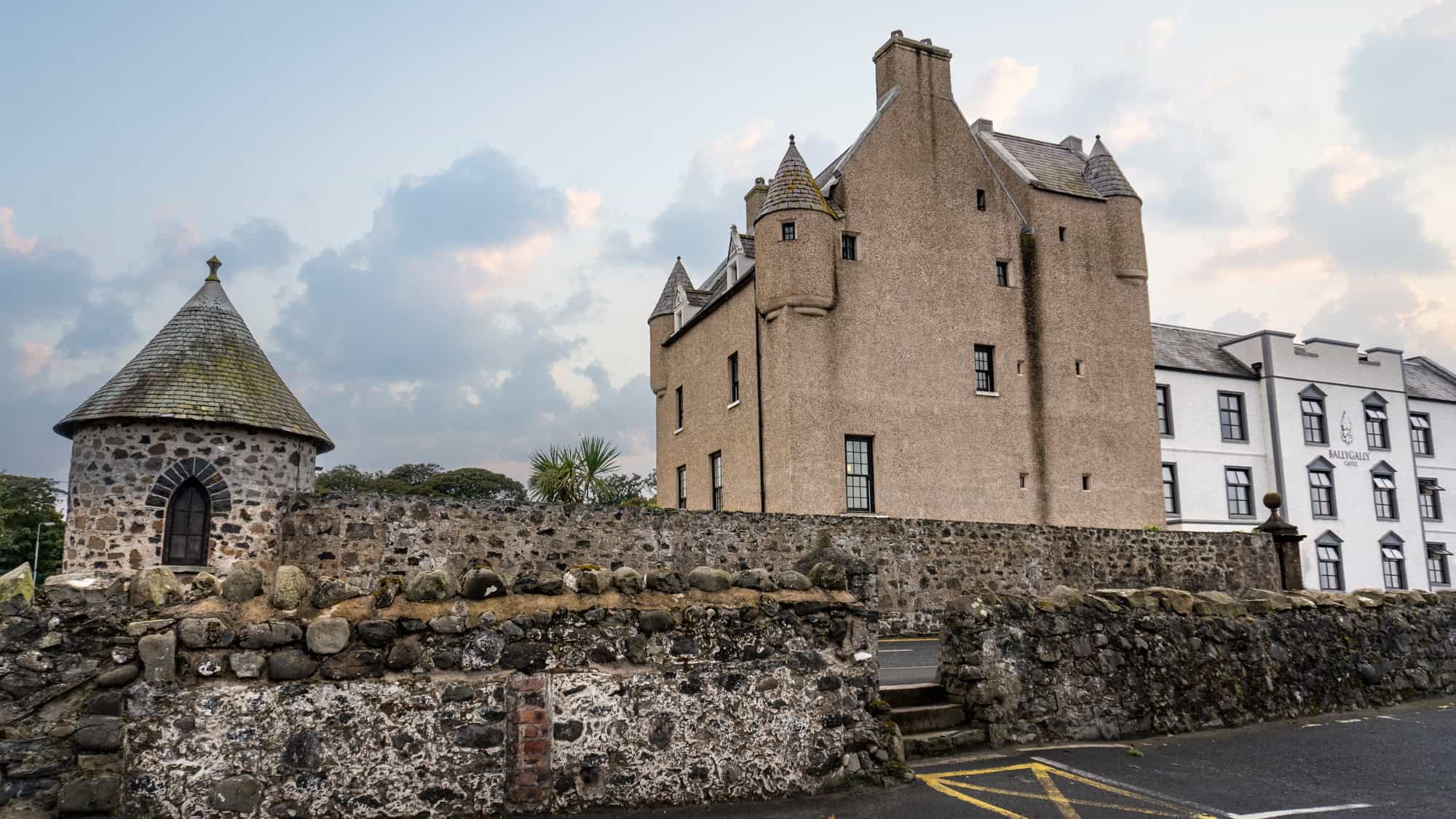 A historic castle with a steeply pitched roof and turrets sits behind a stone wall, with a conical-roofed outbuilding in the foreground. The sky is painted in soft hues as the castle contrasts against the modern hotel beside it.
