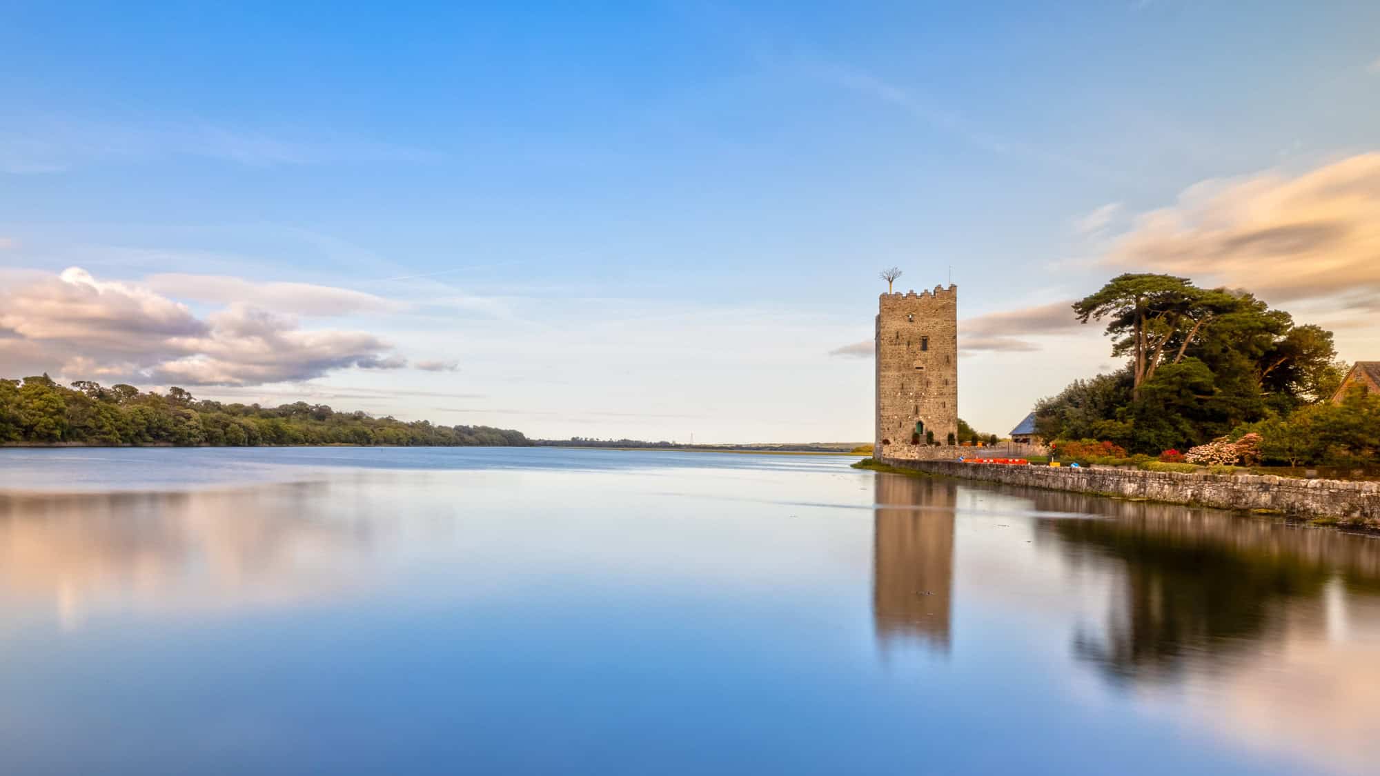 A medieval stone tower stands along the water's edge, reflecting in the calm waters of the river. The sky is painted in soft blues and oranges, with lush greenery surrounding the historic structure.