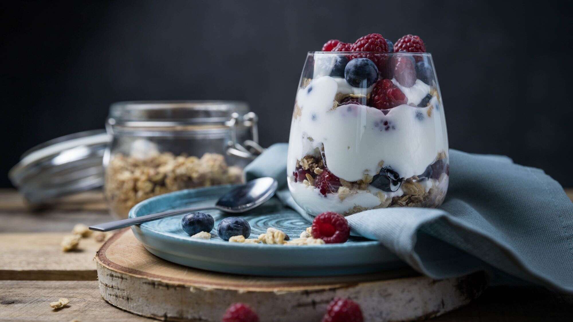 A glass jar filled with layers of creamy yogurt, granola, and mixed berries, topped with a fresh raspberryies and blueberries on a nice blue plate.