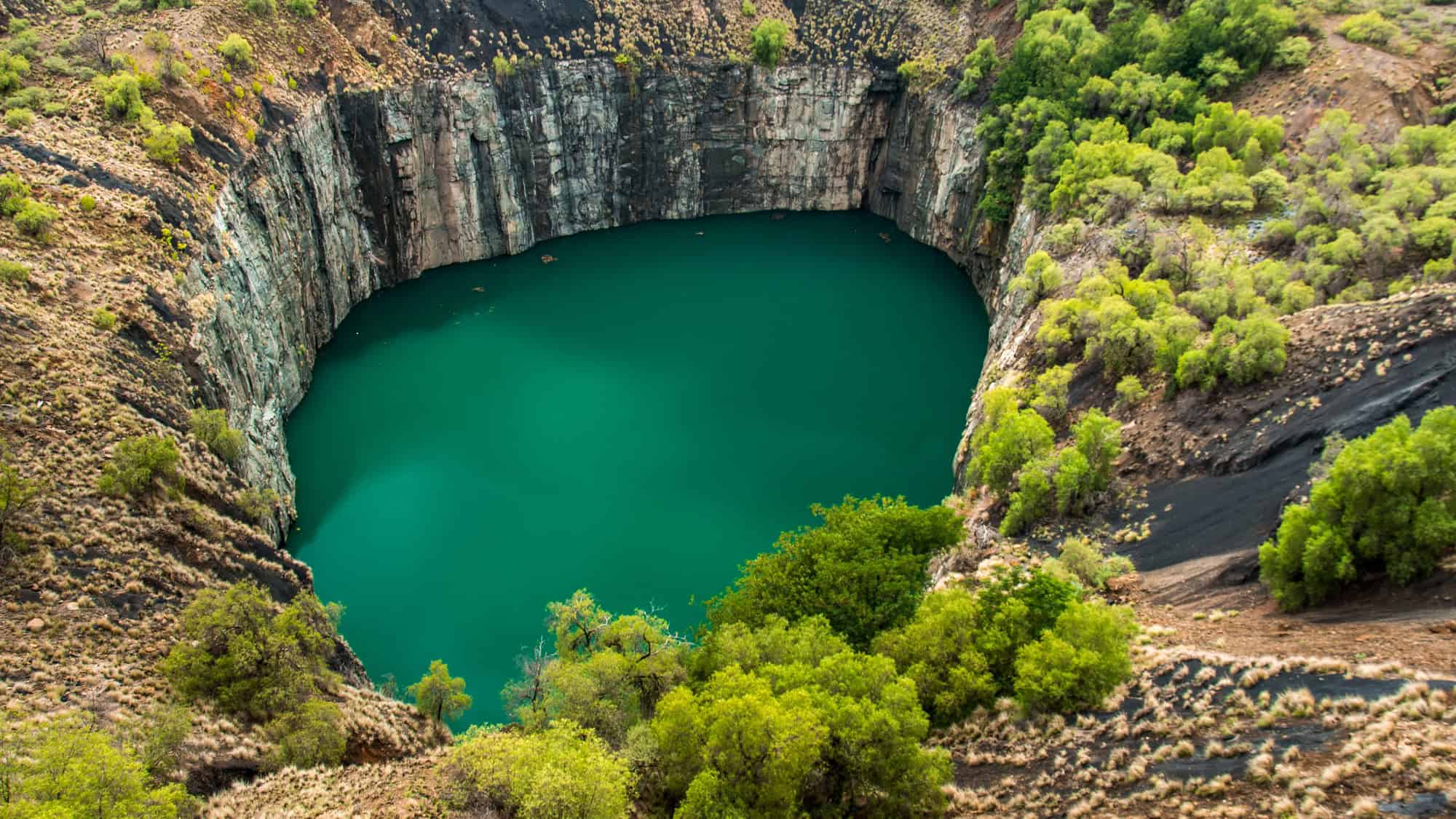A massive circular crater filled with deep turquoise water, surrounded by rugged, rocky cliffs. This historic diamond mine in Kimberley is one of the largest hand-dug excavations in the world.