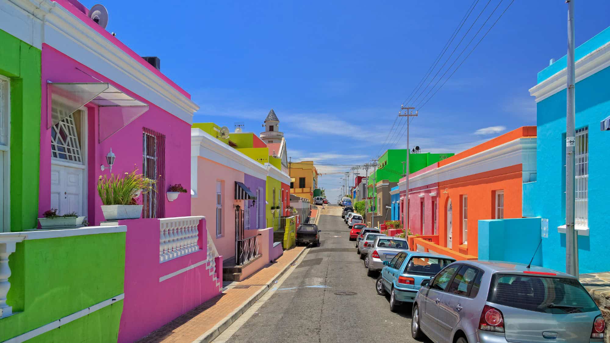 A vibrant street lined with colorful houses in shades of pink, green, blue, and orange under a bright blue sky. Parked cars line the road, adding to the charm of this historic Cape Town neighborhood known for its Cape Malay heritage.