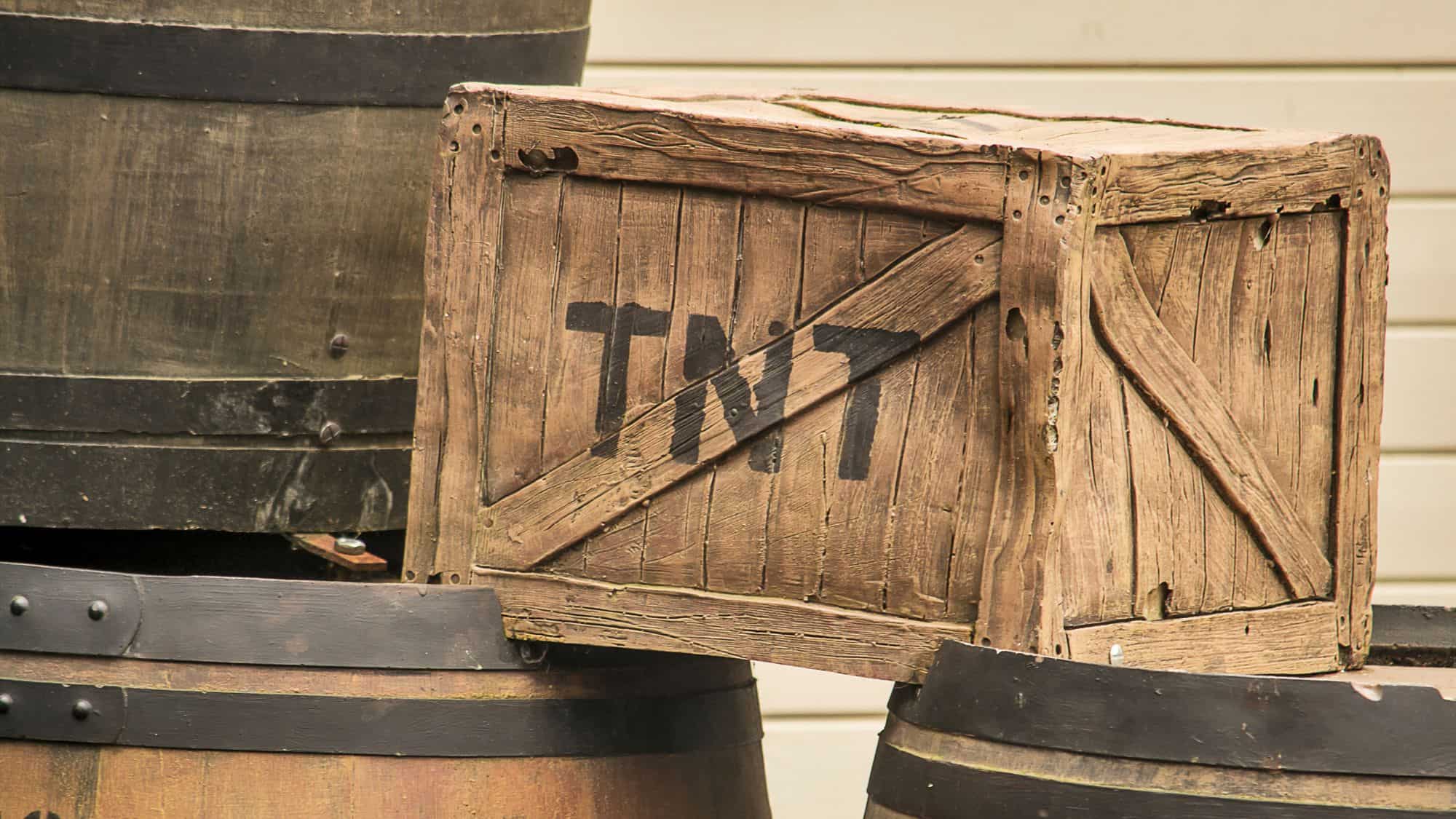 A box of dynamite labeled TNT sits on some wooden barrels outside.