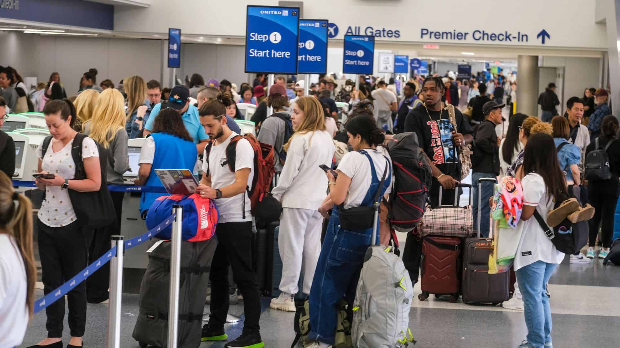 A crowded airport check-in area with travelers standing in long lines, surrounded by luggage and signs indicating "Step 1 Start Here" and "Premier Check-In." The atmosphere is bustling with people checking their phones, reading documents, and waiting.