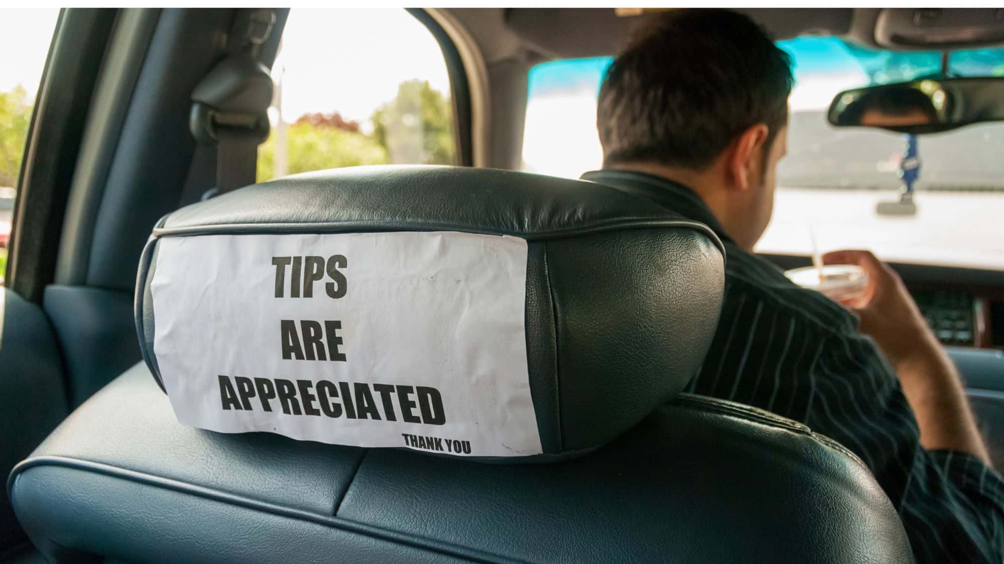 A close-up of the back of a taxi driver's seat with a paper sign taped to it that reads "TIPS ARE APPRECIATED THANK YOU." The driver is visible in the background, holding a drink while driving.