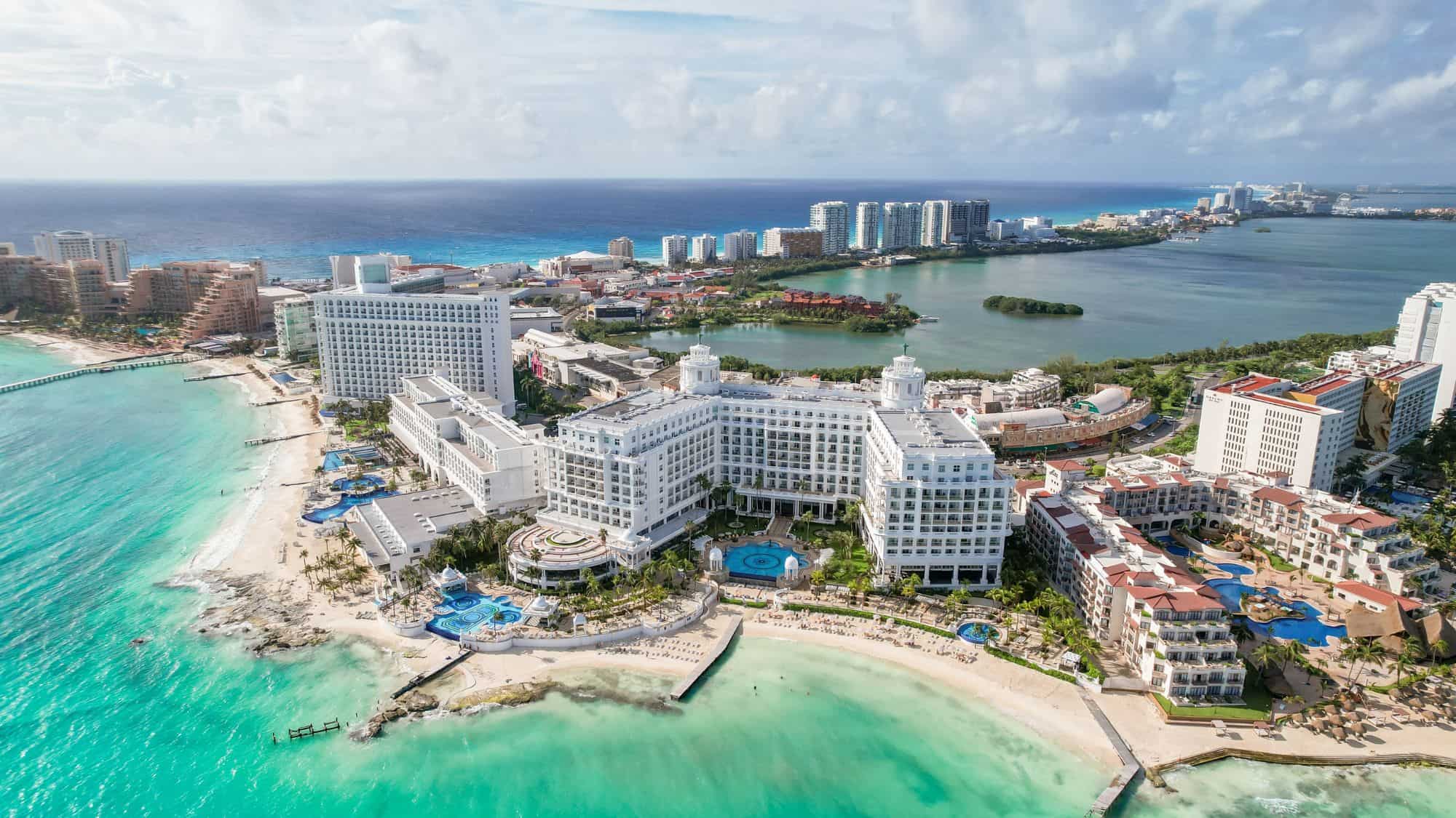 An aerial view of Cancun’s hotel zone, showcasing luxurious white beachfront resorts surrounded by turquoise waters. The coastline extends into the distance, bordered by lush green islands and the deep blue Caribbean Sea.