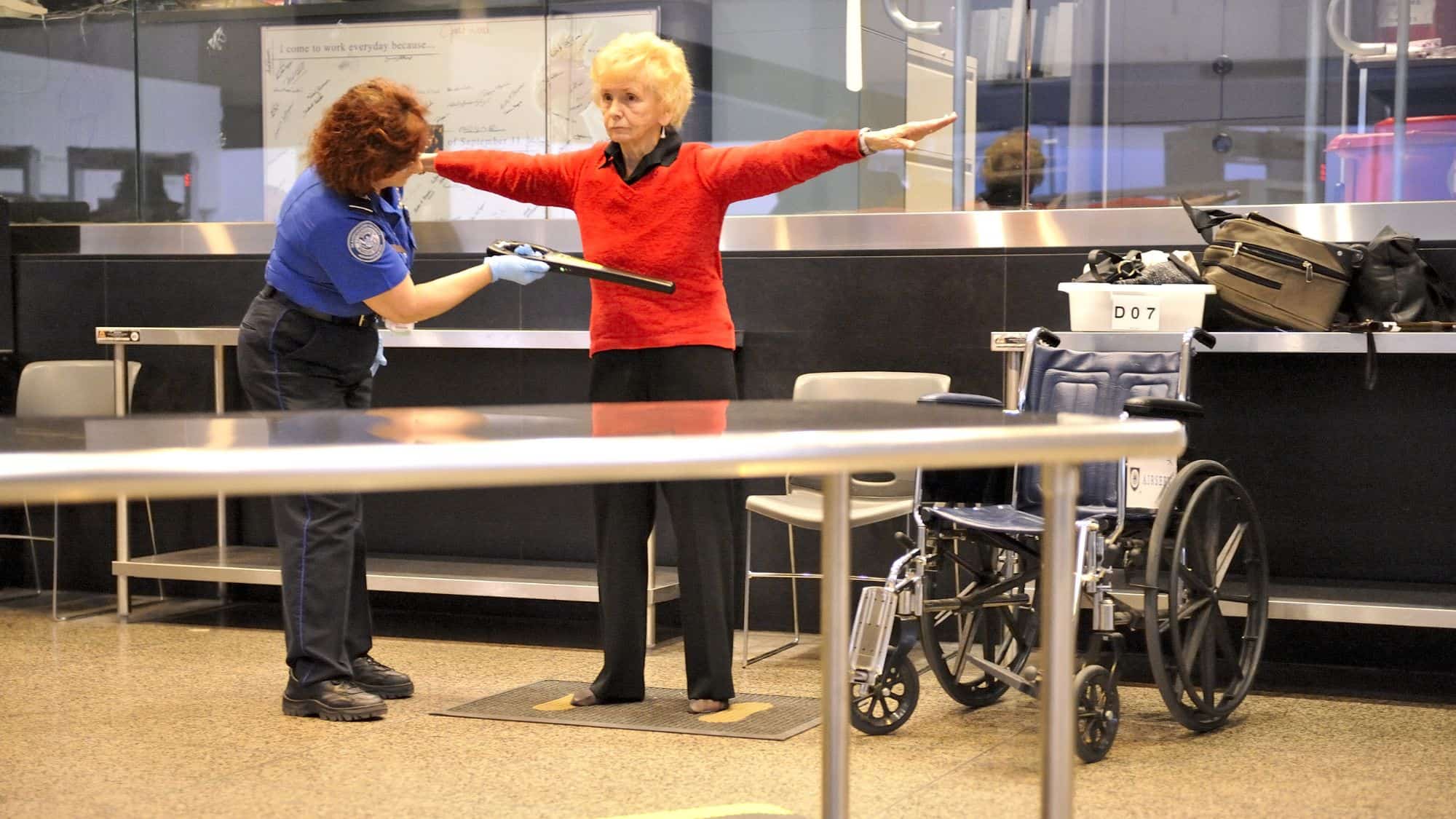 A TSA officer in a blue uniform conducts a security screening on an elderly woman wearing a red sweater, who stands with her arms outstretched. A wheelchair and personal belongings are visible in the background at the airport security checkpoint.