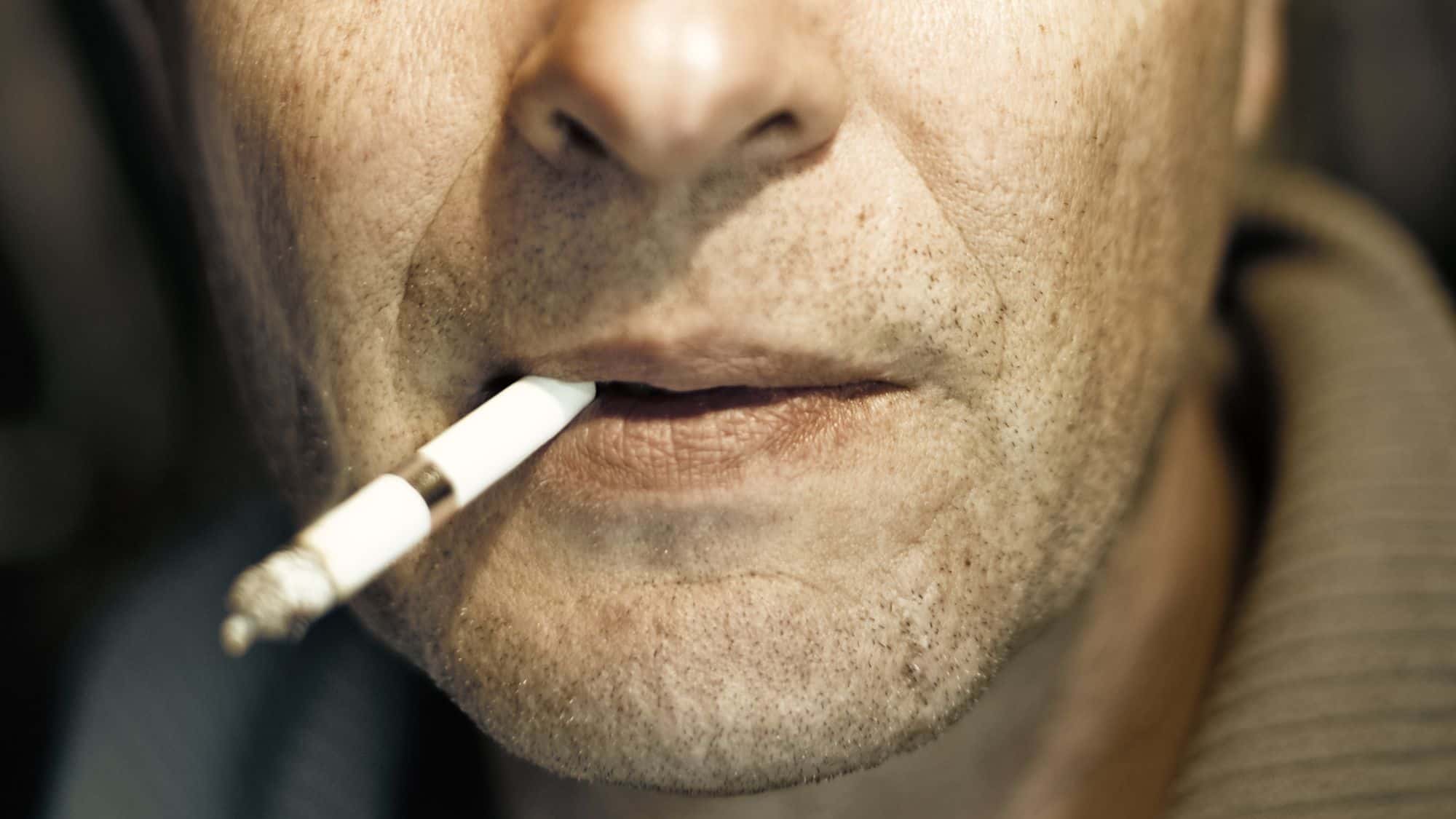 A close-up shot of a man's face with a cigarette between his lips. His rough skin and stubble are highlighted by moody lighting.