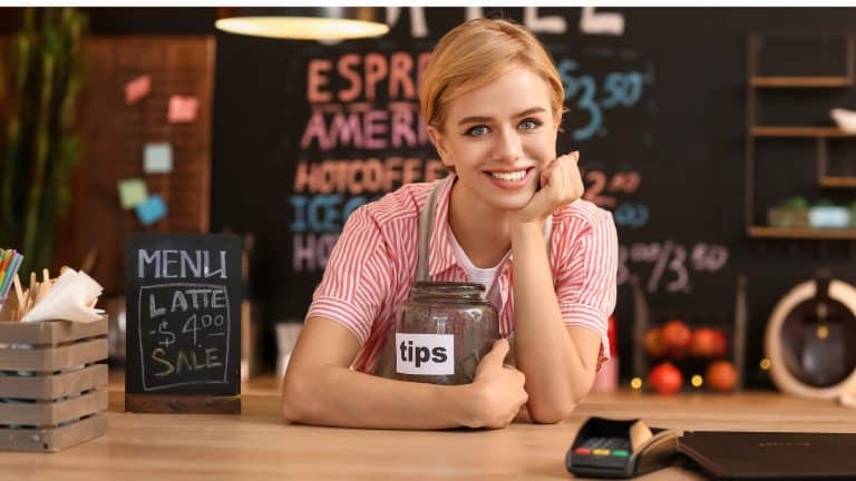 A smiling barista leans on a counter, resting her chin on her hand while holding a glass tip jar labeled "TIPS." Behind her is a café menu board with colorful chalk writing.