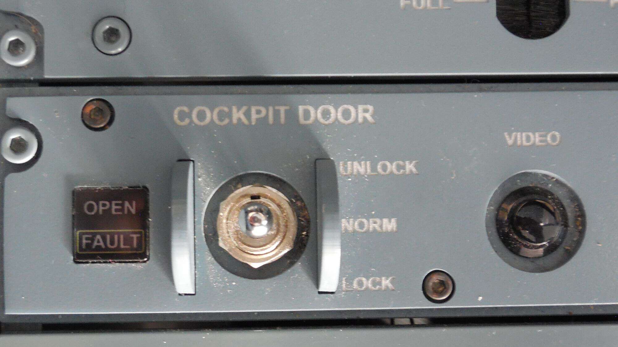 A close-up of an aircraft's cockpit door control panel, showing a keyhole, lock and unlock switches, a fault indicator, and a video monitor knob. The panel appears slightly worn from frequent use.