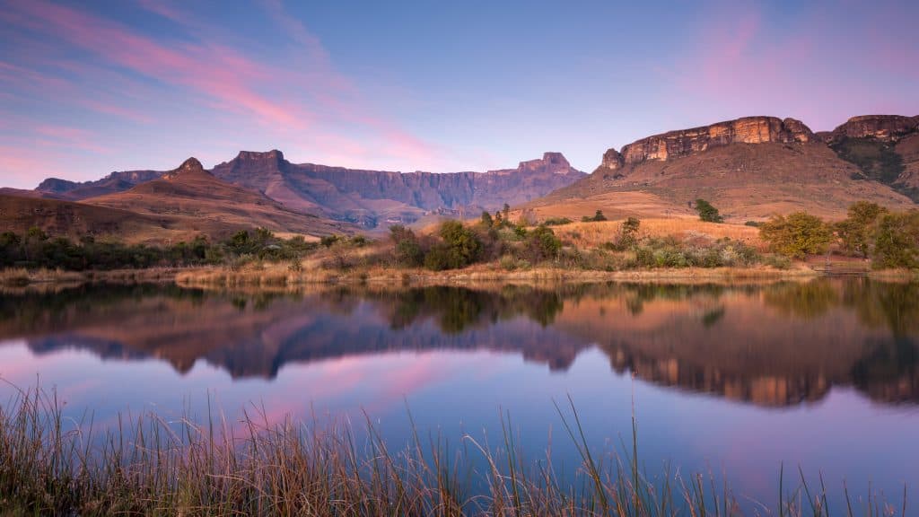A serene lake reflecting the towering Drakensberg peaks under a soft, pastel-colored sky at dusk. The still water mirrors the golden hues of the mountains, creating a peaceful and picturesque setting.
