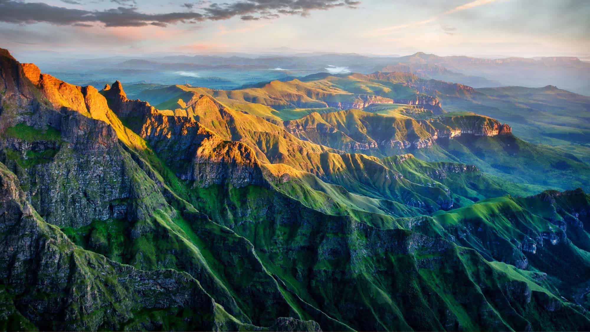 A stunning aerial view of the Drakensberg mountain range, bathed in warm golden light. The jagged green peaks and deep valleys stretch as far as the eye can see.