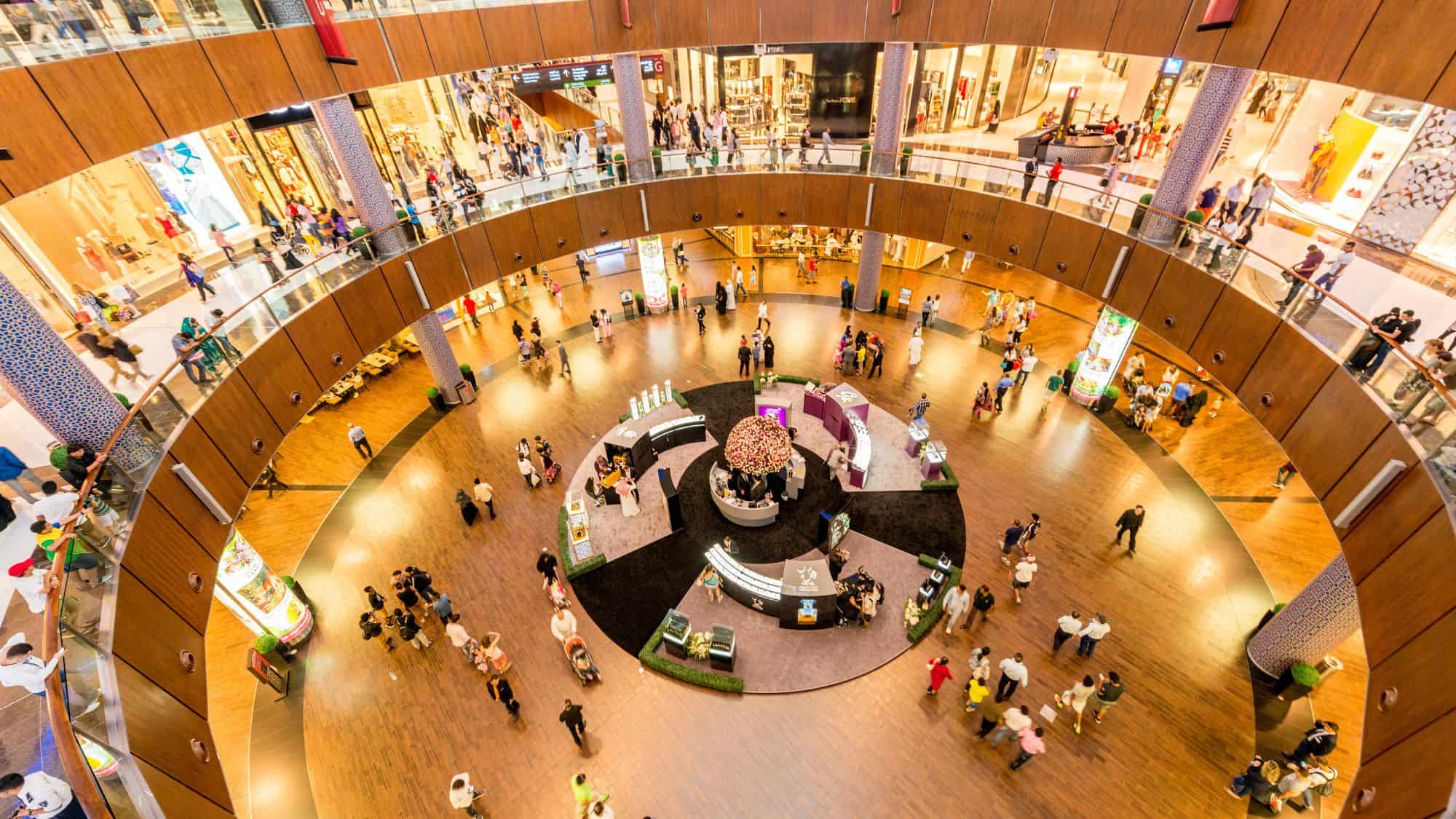 A vast, circular atrium inside Dubai Mall, with people walking along the glossy wooden floors and luxury brand stores lining the upper levels. The modern, well-lit space features a central seating area surrounded by high-end shops.