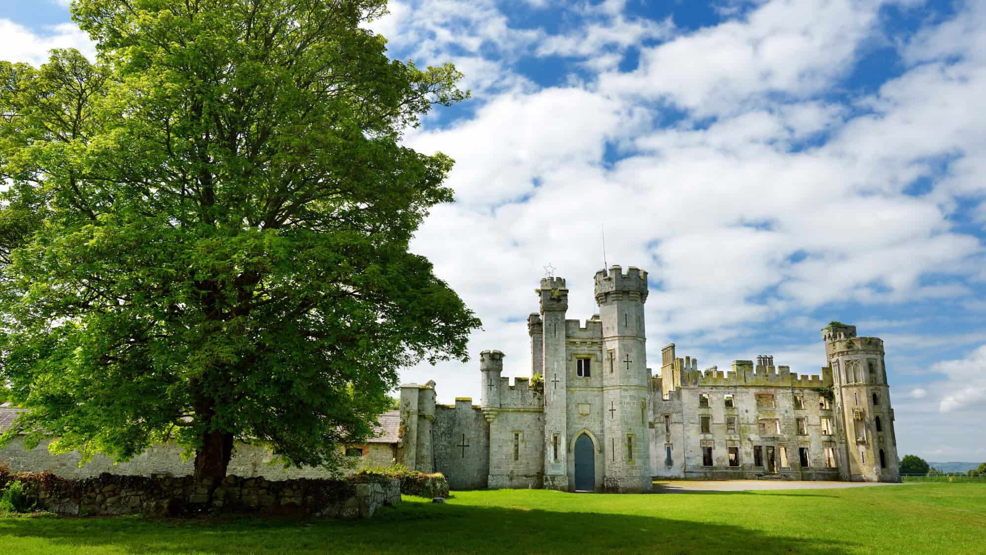 The ruins of an elaborate gothic revival mansion with multiple turrets and an overgrown courtyard. The structure stands partially intact, its once-grand halls now open to the elements.