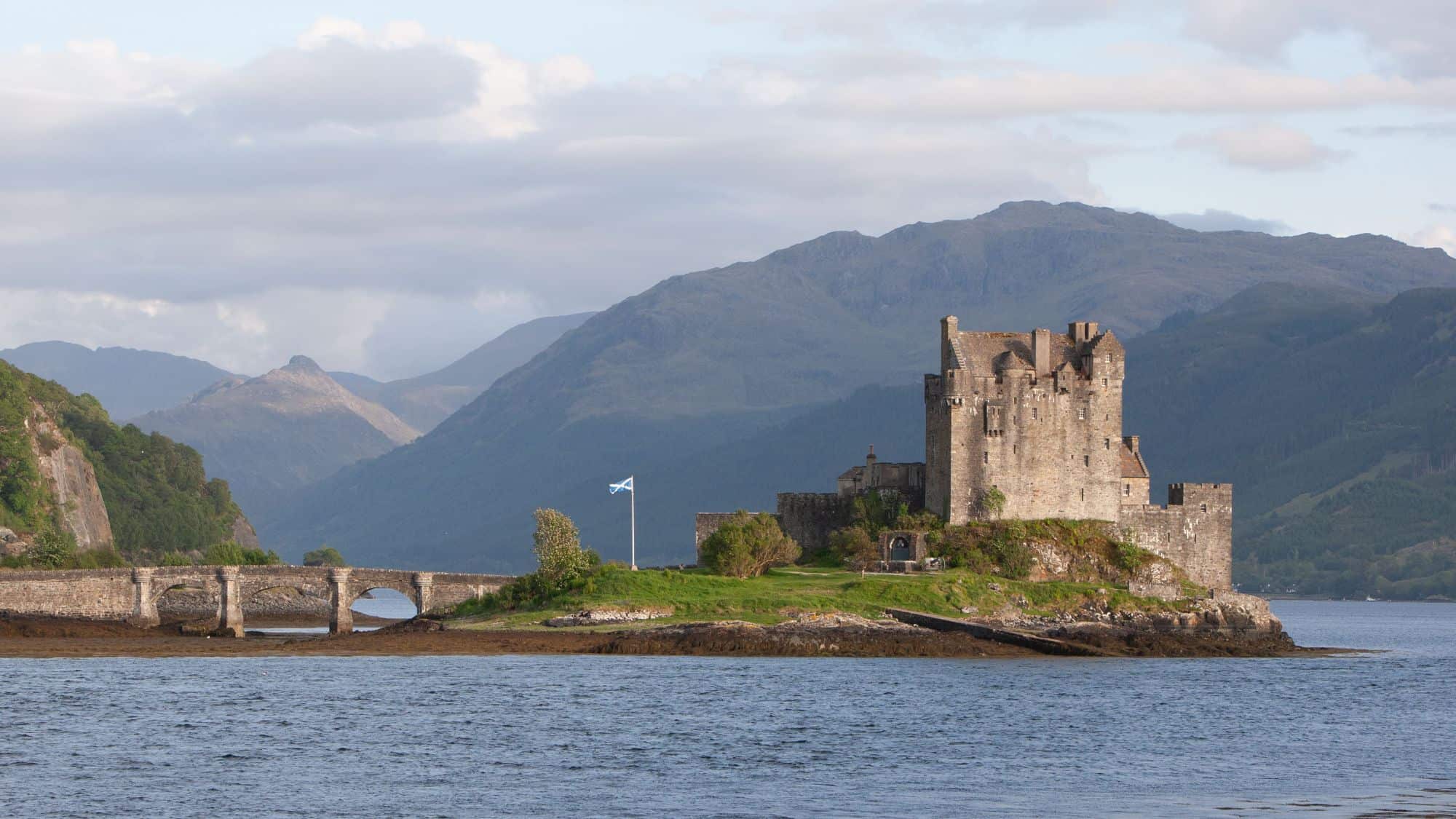 Eilean Donan Castle stands on a small island where three lochs meet, connected by an old stone bridge. The castle is framed by rolling hills and calm waters, with the Scottish flag flying nearby.