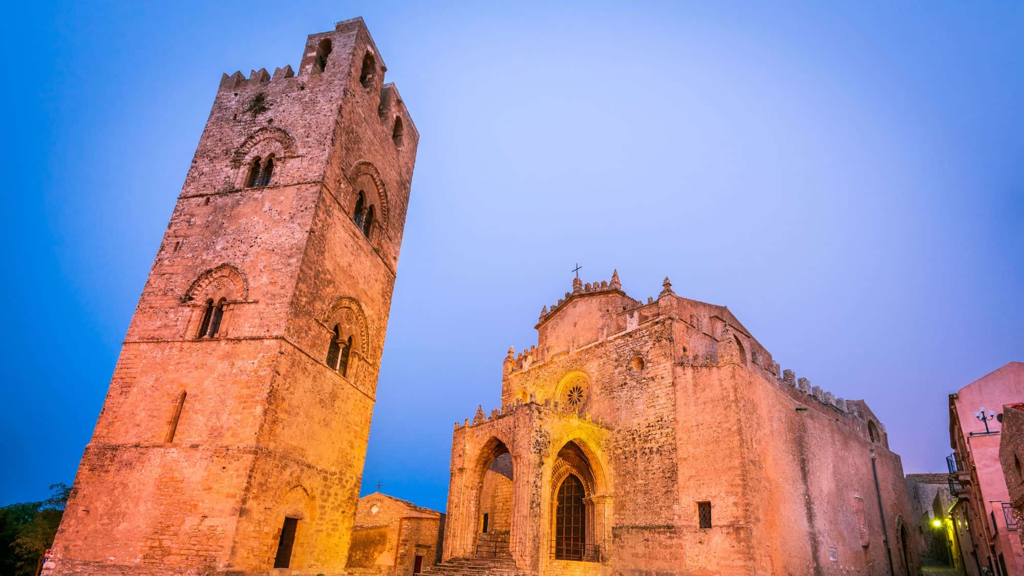 The historic cathedral of Erice, Sicily, with its imposing medieval bell tower and gothic-style façade glowing under the soft evening light. The ancient stonework and pointed arches give it a timeless and mystical charm.
