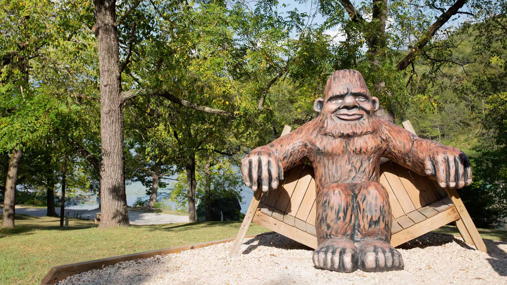 A large wooden statue of Bigfoot sitting on a rustic bench in a park surrounded by lush green trees. The scene is set near a lake, with picnic benches and a gravel path nearby.