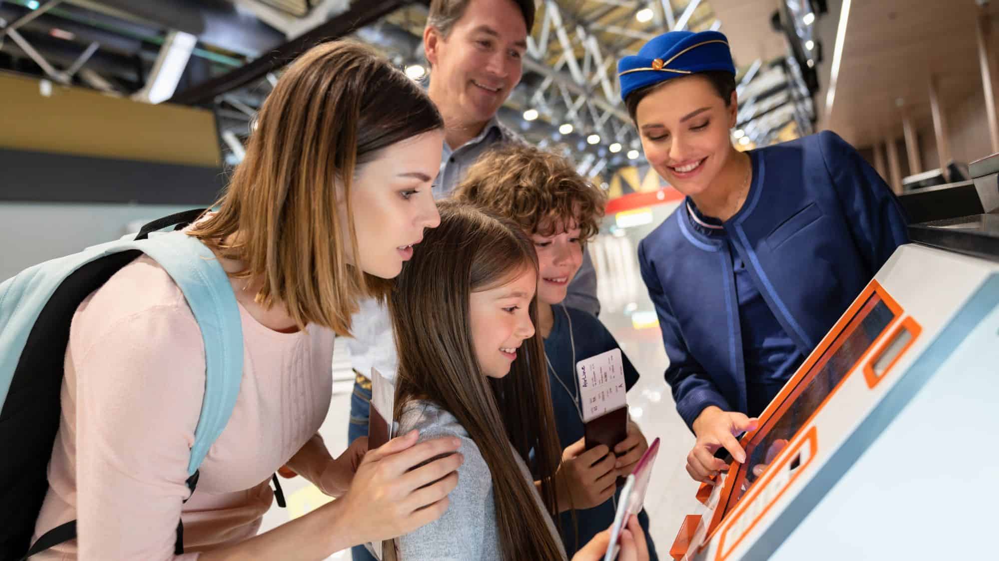 A family of four checks in at an airport kiosk with the help of a friendly airline staff member in a navy blue uniform. The mother, father, and two children smile as they receive travel assistance.