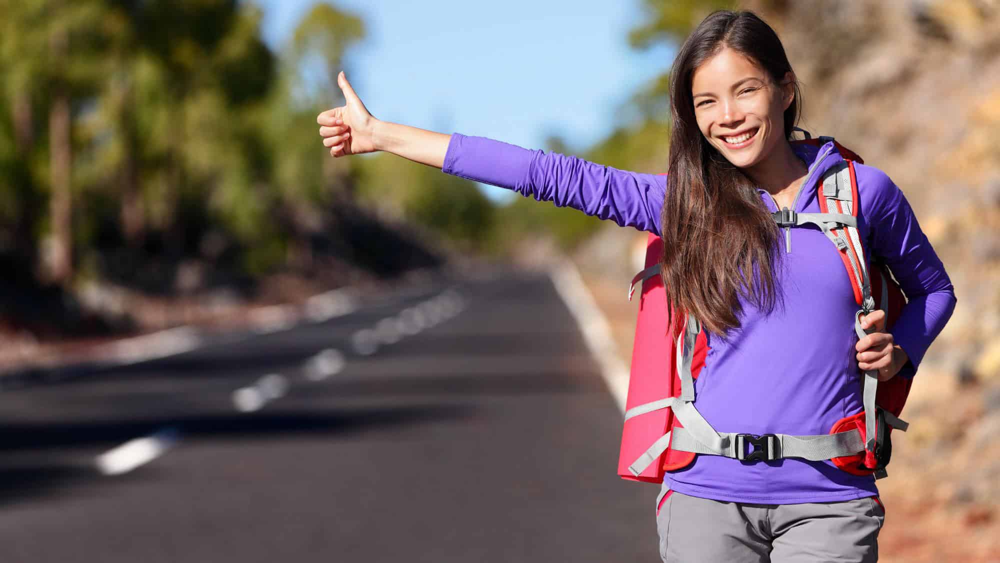 A young woman with a bright smile and a red hiking backpack sticks out her thumb on an empty road, hoping to catch a ride. The scenery around her is filled with trees and hills.