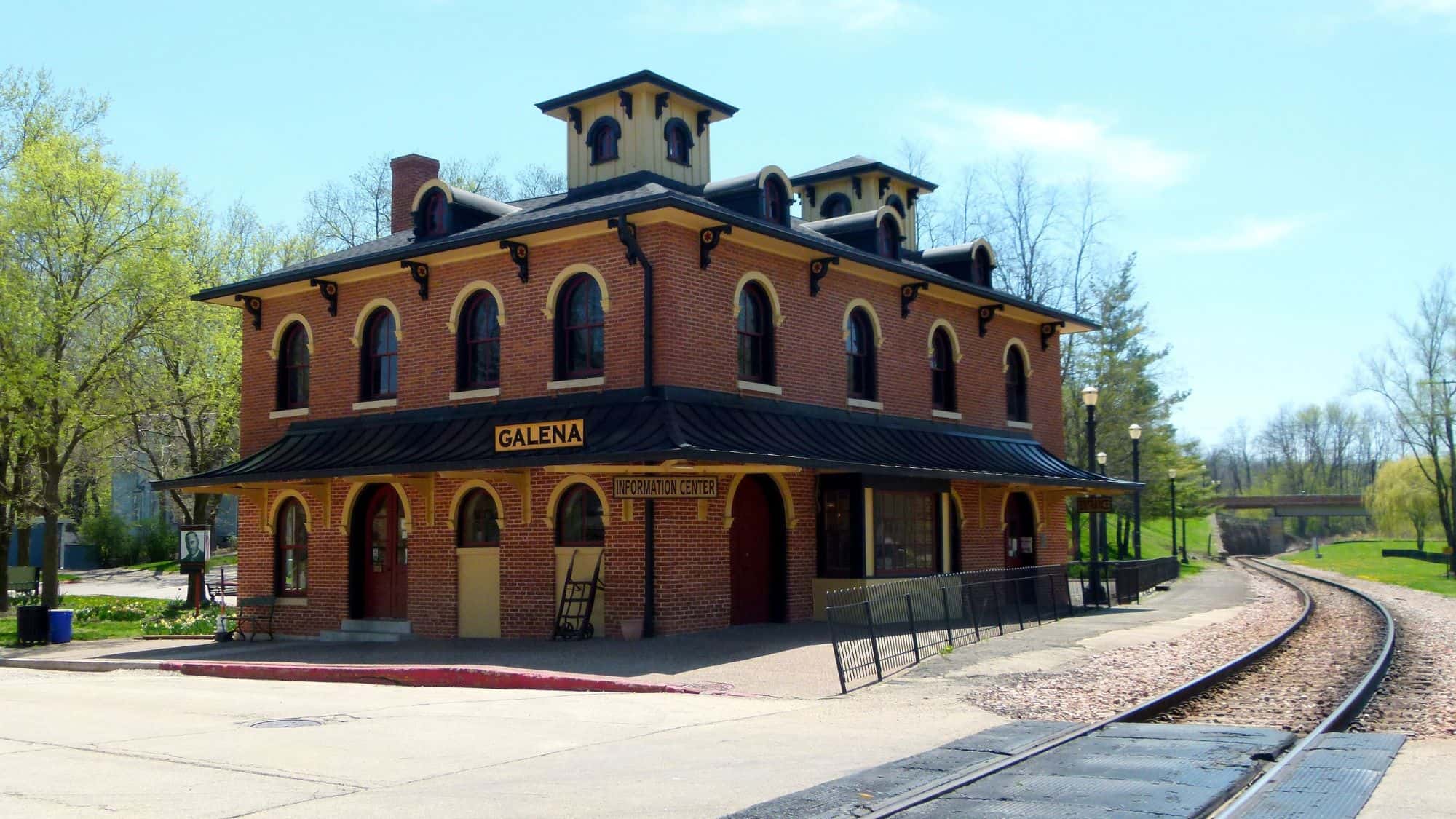 A historic red-brick train station in Galena, Illinois, now serving as an information center. The building features arched windows, black trim, and signage, with a railroad track curving into the distance alongside it.
