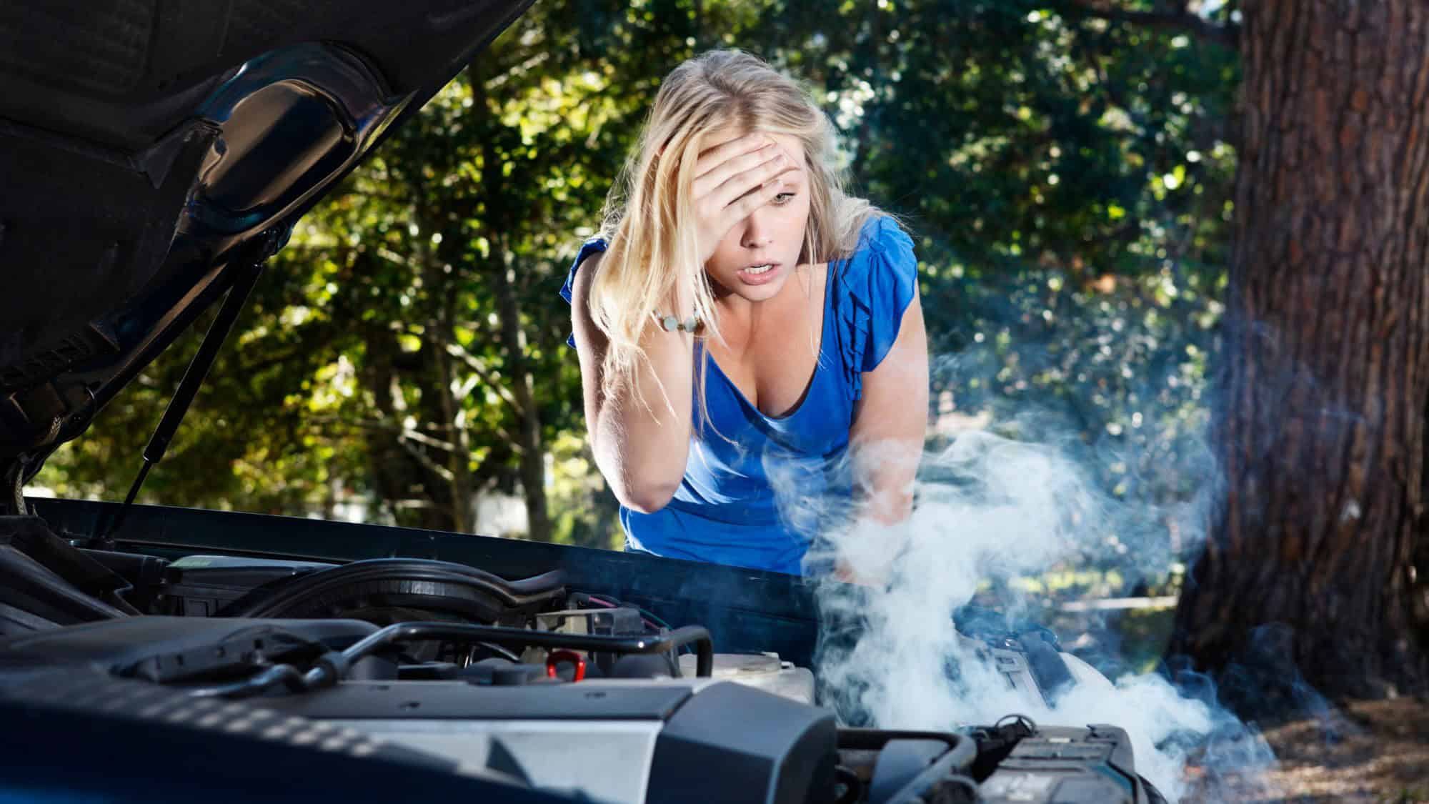 A frustrated young woman in a blue shirt stands beside a broken-down car with smoke rising from the engine. She holds her head in exasperation as she looks at the damage.