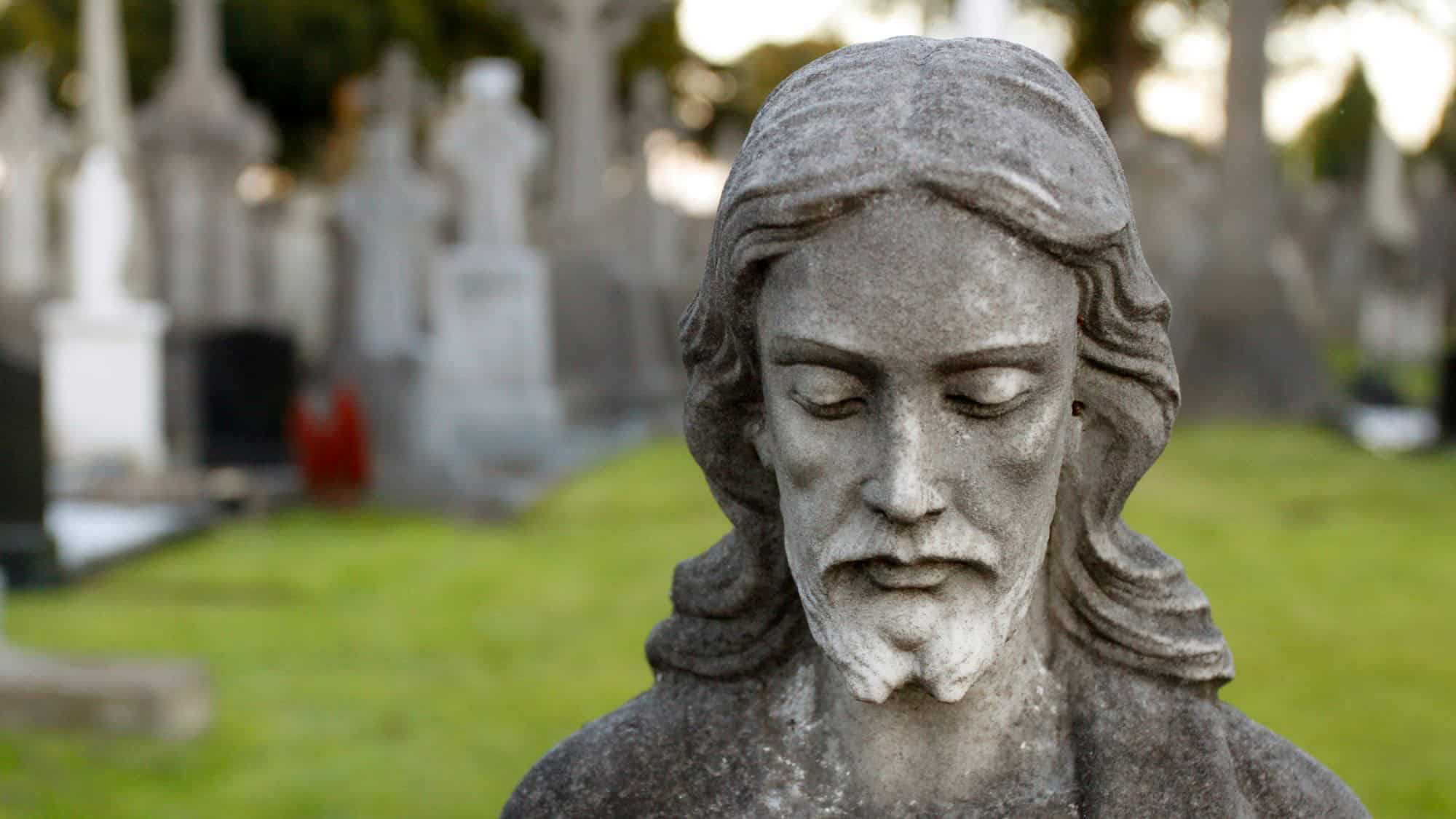 A weathered stone statue of Jesus Christ is seen in the foreground, looking downward with a solemn expression. Behind it, rows of tombstones stretch across a green cemetery, creating an eerie yet peaceful atmosphere.
