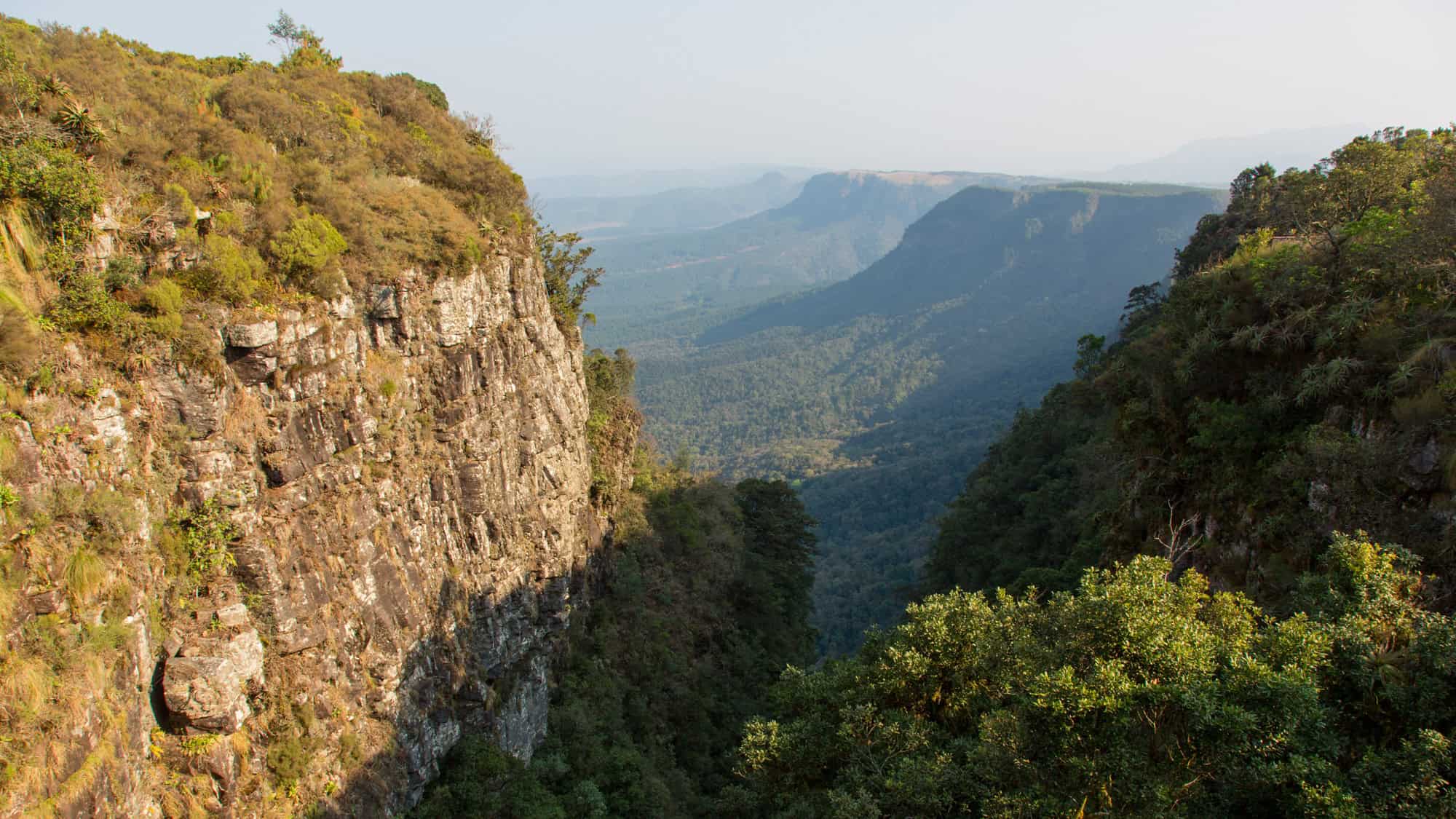 A dramatic landscape showcasing a steep cliffside covered in lush vegetation, overlooking a vast valley. The distant mountains fade into the mist, creating an ethereal effect.