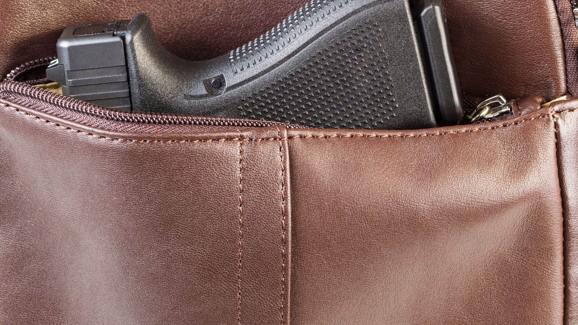 A black handgun partially concealed inside a brown leather zippered bag. The close-up shot emphasizes the firearm’s handle and texture.