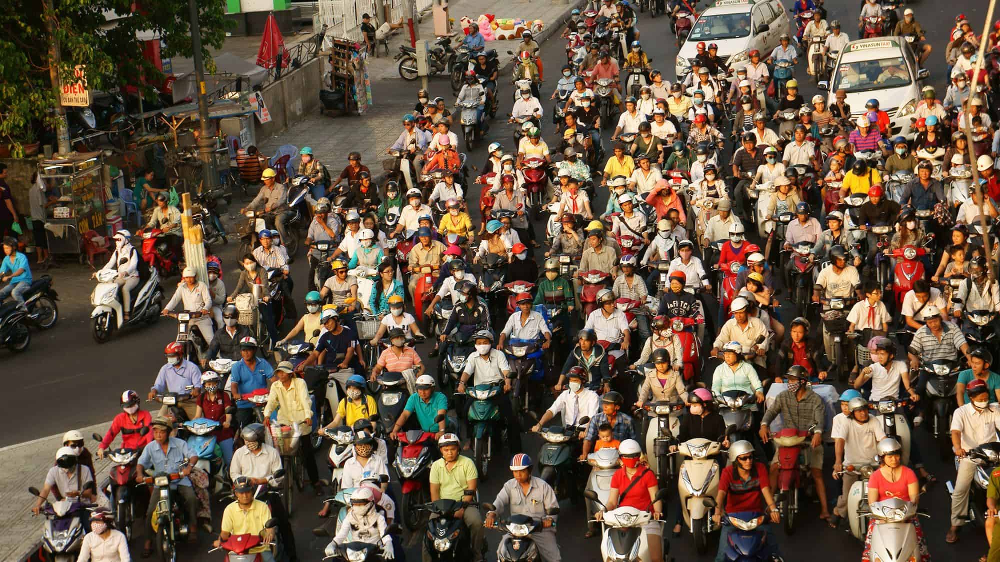 A chaotic scene of a street in Hanoi, Vietnam, packed with motorbikes as riders, many wearing helmets and masks, navigate through dense traffic. The crowd of scooters dominates the road, with pedestrians and street vendors visible on the sides.