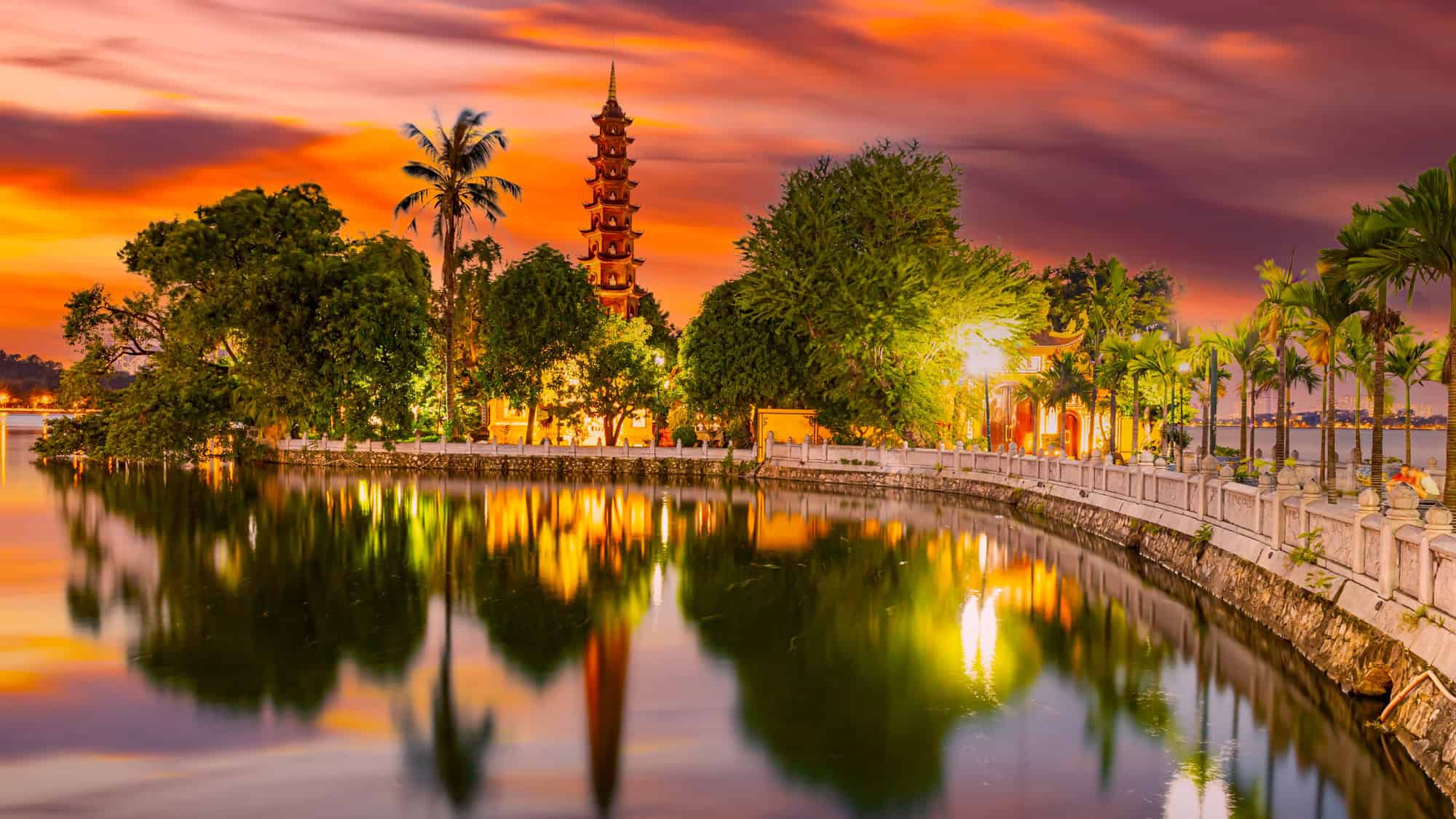 The Tran Quoc Pagoda in Hanoi, Vietnam, reflected in the still waters of West Lake during a vibrant sunset, surrounded by lush greenery and palm trees.