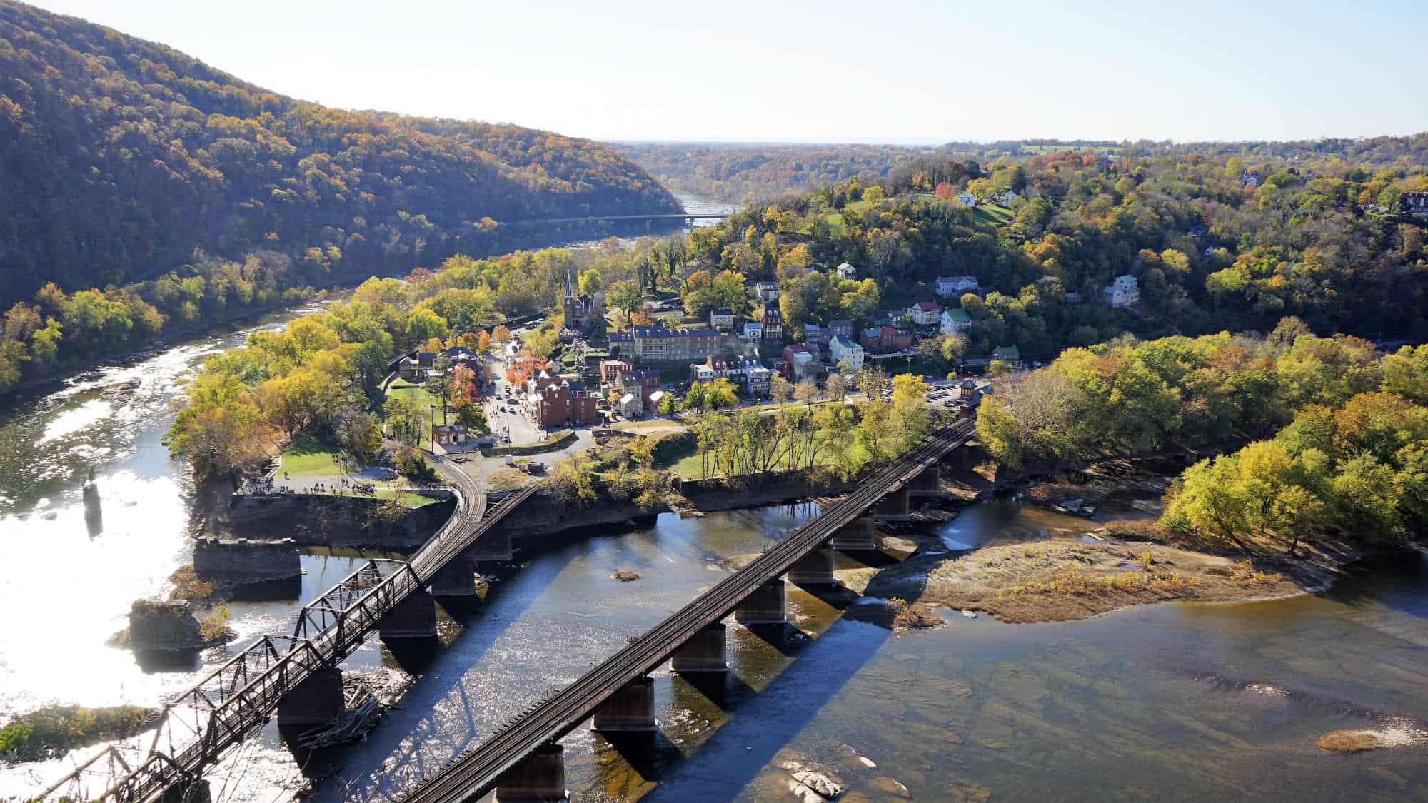 An aerial view of Harpers Ferry, West Virginia, where the Potomac and Shenandoah Rivers meet. The small town is nestled among rolling hills covered in autumn foliage, with a historic railway bridge crossing the river.