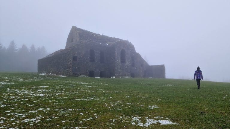 A ruined stone structure sits atop a grassy hill, partially shrouded in mist. A lone figure in a colorful jacket walks toward the abandoned building, adding an eerie and mysterious element to the scene.