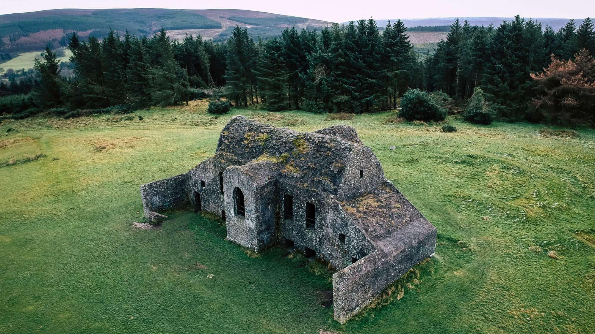 A crumbling stone ruin sits isolated on a grassy hillside, surrounded by dark evergreen trees. Its moss-covered walls and eerie openings suggest a mysterious past.