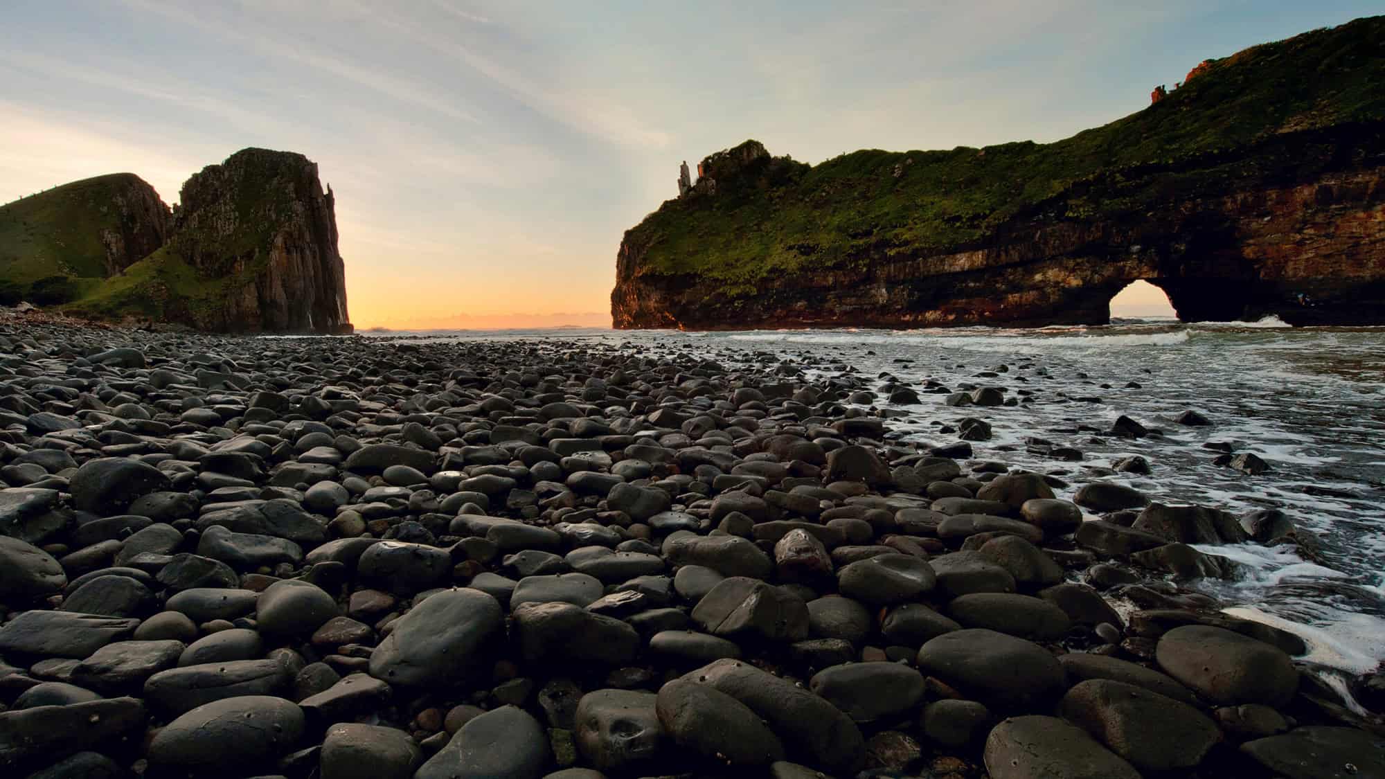 A dramatic rock formation on the Wild Coast, with a naturally carved arch standing against the crashing waves. The pebble-covered shoreline and warm sunset hues add to the mystique.