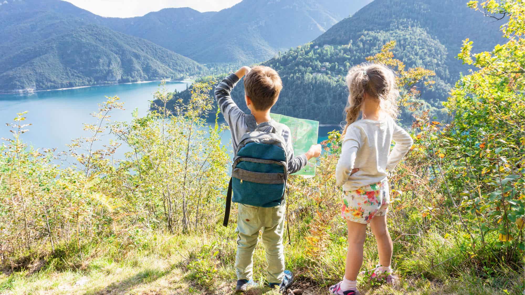 Two children stand on a hillside overlooking a scenic lake, surrounded by lush greenery. The boy, wearing a backpack, holds a map while the girl, in floral shorts, gazes into the distance.