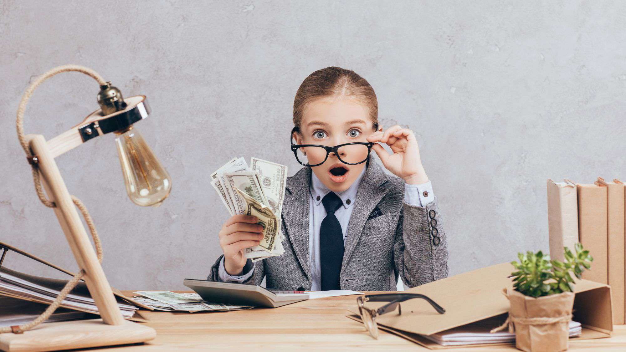 A young girl dressed in a business suit holds a stack of U.S. dollar bills in one hand and adjusts her oversized glasses with the other. Her surprised expression adds humor to the scene, as she sits at a desk with office supplies.