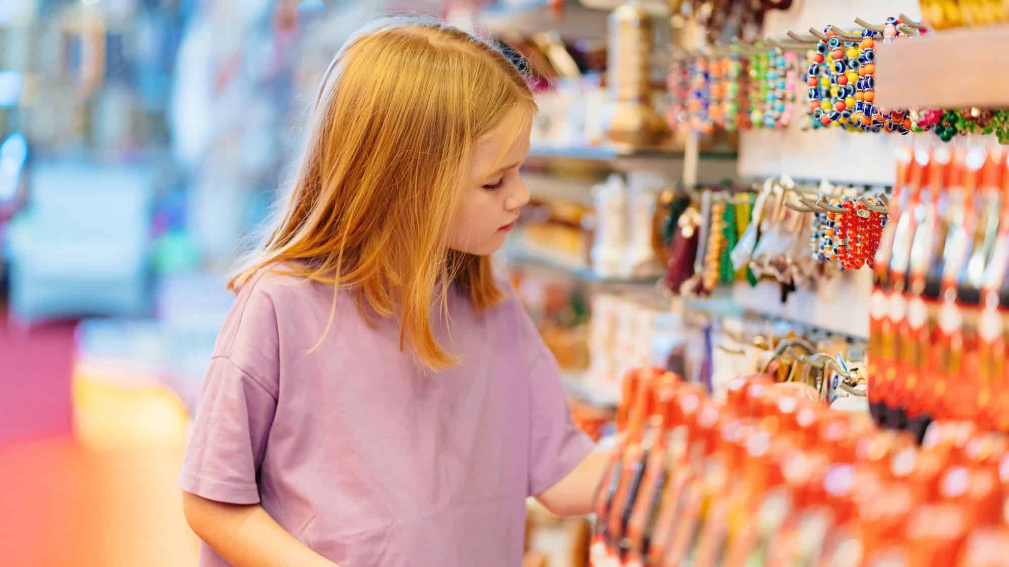 A young girl with blonde hair and a purple shirt browses a souvenir shop filled with colorful bracelets, trinkets, and bottled goods. She looks at the items on display with curiosity in a well-lit, vibrant store.