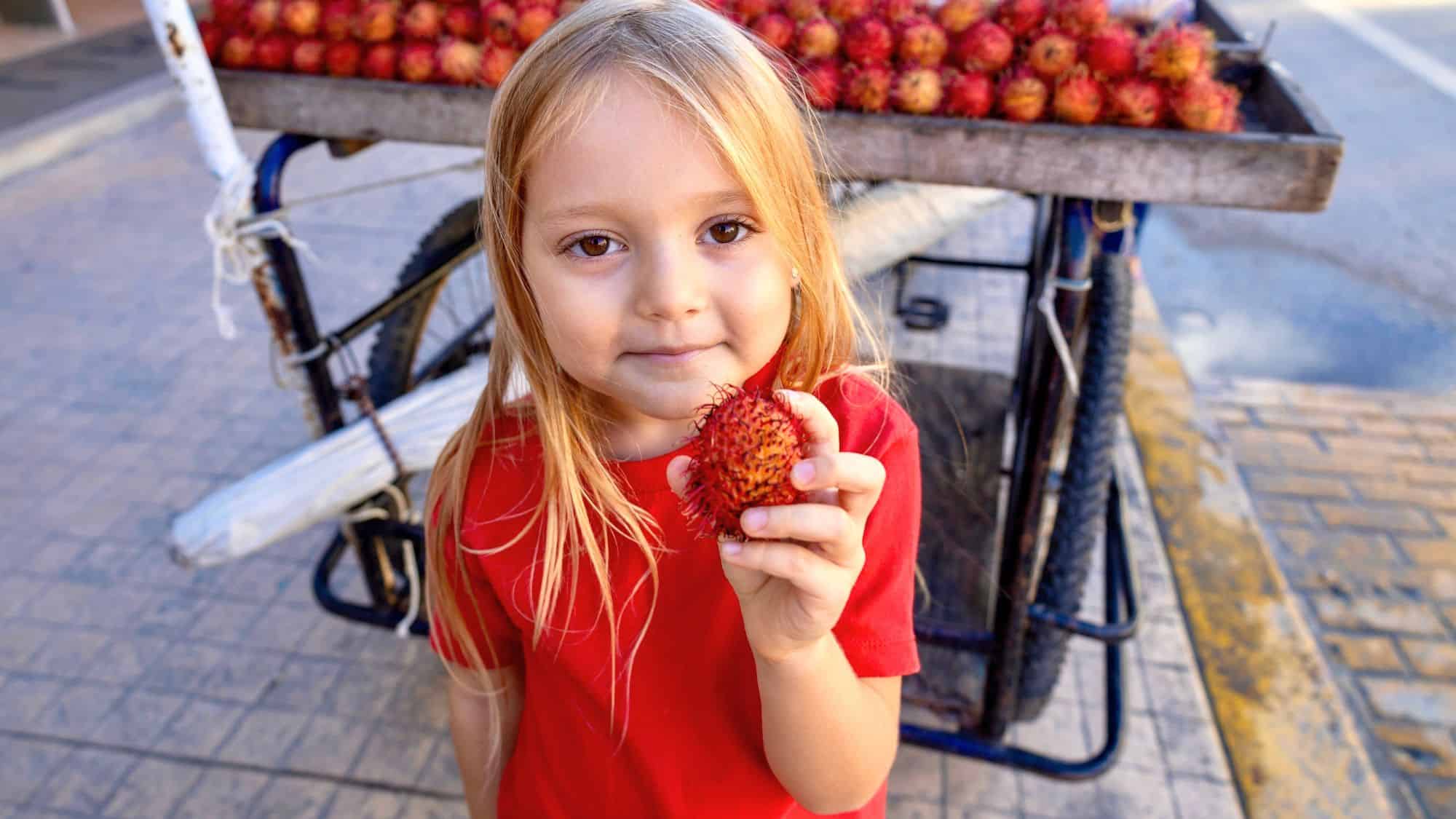 A young blonde girl in a red shirt holds a rambutan fruit close to the camera, smiling slightly. Behind her, a bicycle cart filled with fresh rambutan is parked on the street.