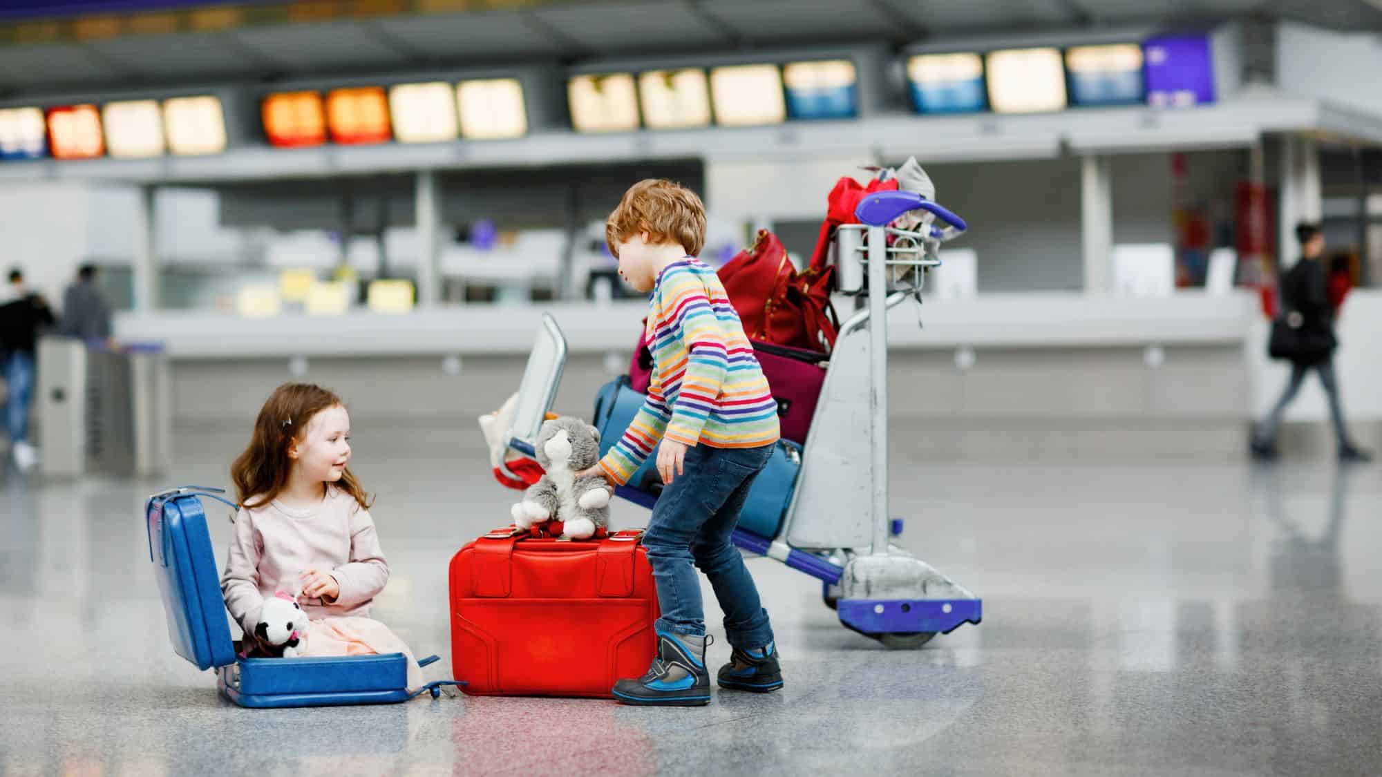 Two young children play with luggage in an airport terminal. A girl sits inside an open suitcase holding a stuffed animal, while a boy in a striped sweater places another plush toy on top of a red suitcase.