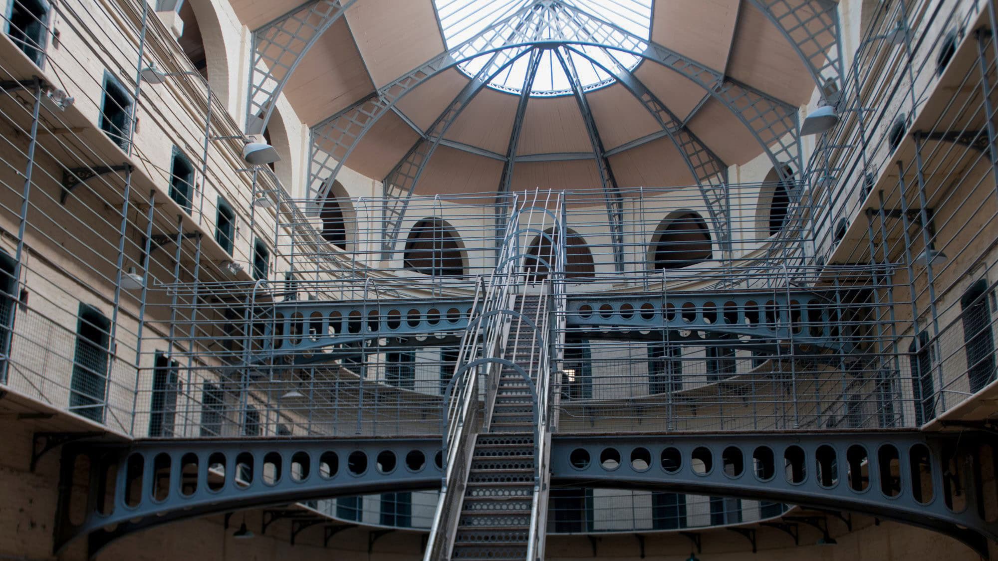 The interior of an old prison with multiple levels of metal railings and arched doorways, all centered around a grand staircase. The domed ceiling lets in natural light, illuminating the cold, structured space.