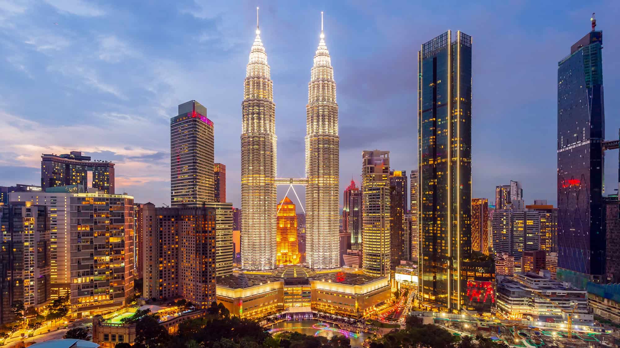A stunning evening cityscape of Kuala Lumpur, Malaysia, featuring the illuminated Petronas Twin Towers standing tall among the surrounding skyscrapers. The city's lights reflect vibrantly against the twilight sky.
