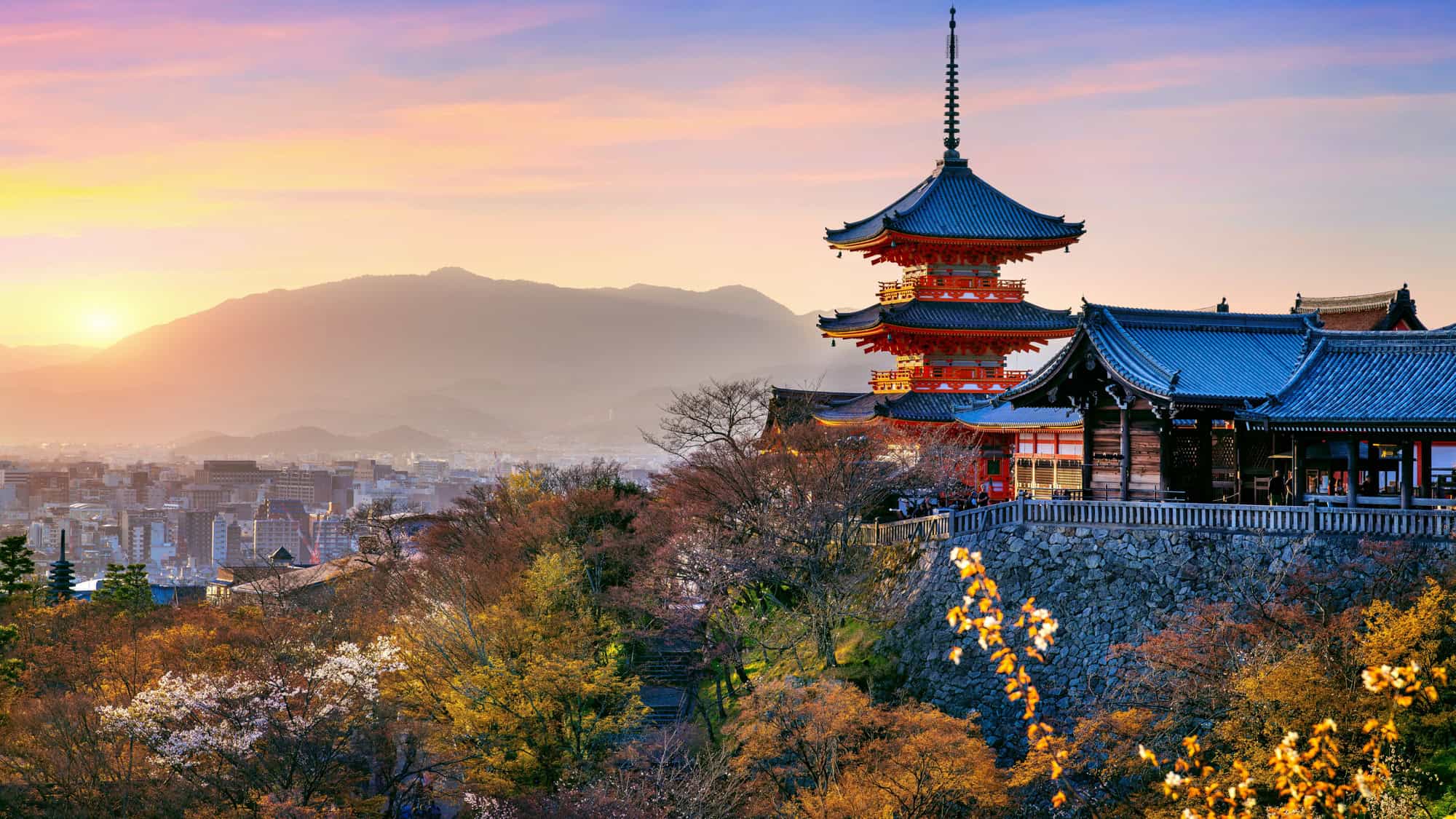 The Kiyomizu-dera Temple in Kyoto, Japan, stands elegantly against a backdrop of autumn foliage and a glowing sunset over the city and distant mountains.