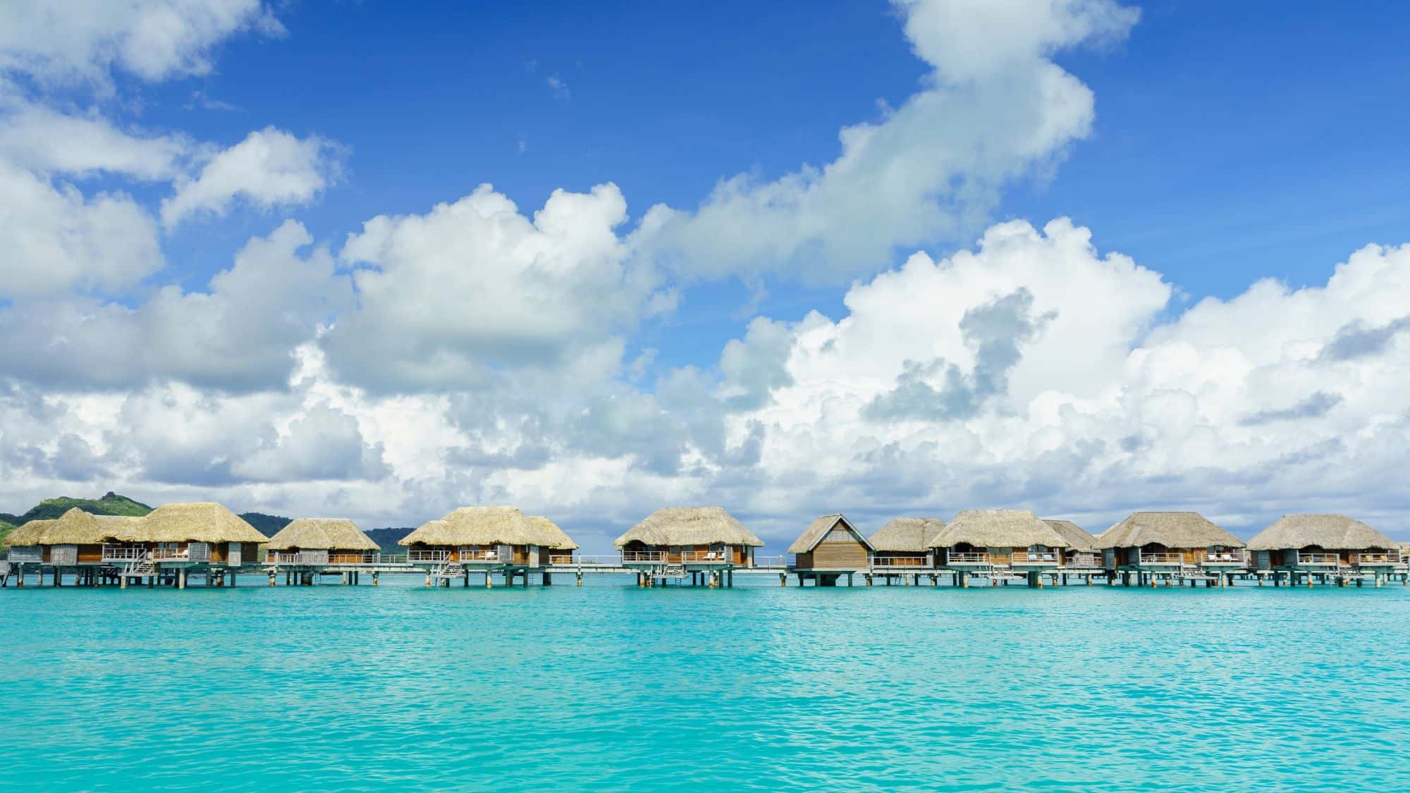 Thatched-roof bungalows stretch across crystal-clear water with lush green hills in the background. The vibrant blue sky is dotted with fluffy white clouds, completing the perfect tropical scene.