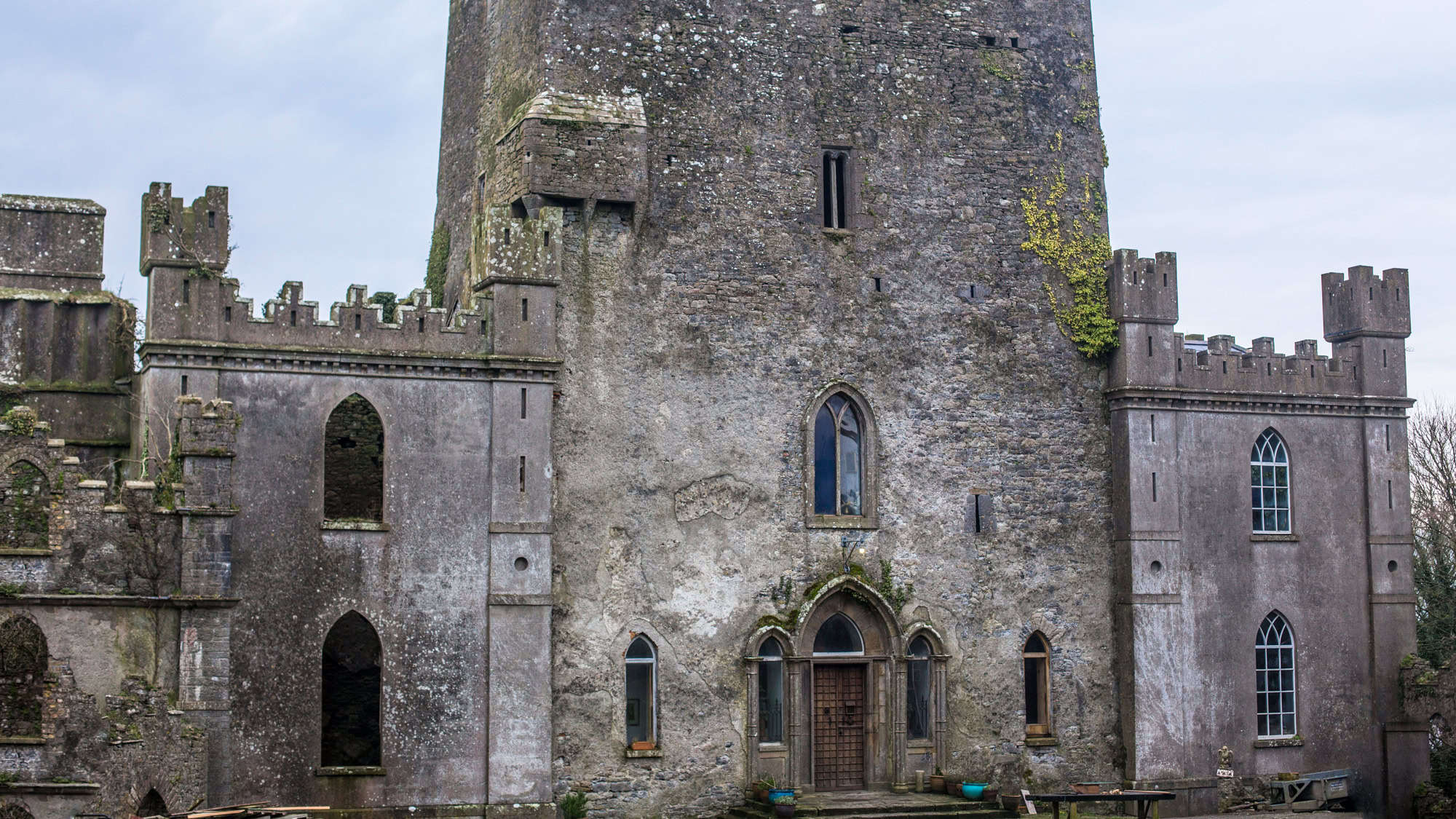 A weathered medieval castle with a rugged stone facade, featuring gothic arched windows and battlements. Patches of ivy climb the walls, hinting at its long and eerie history.