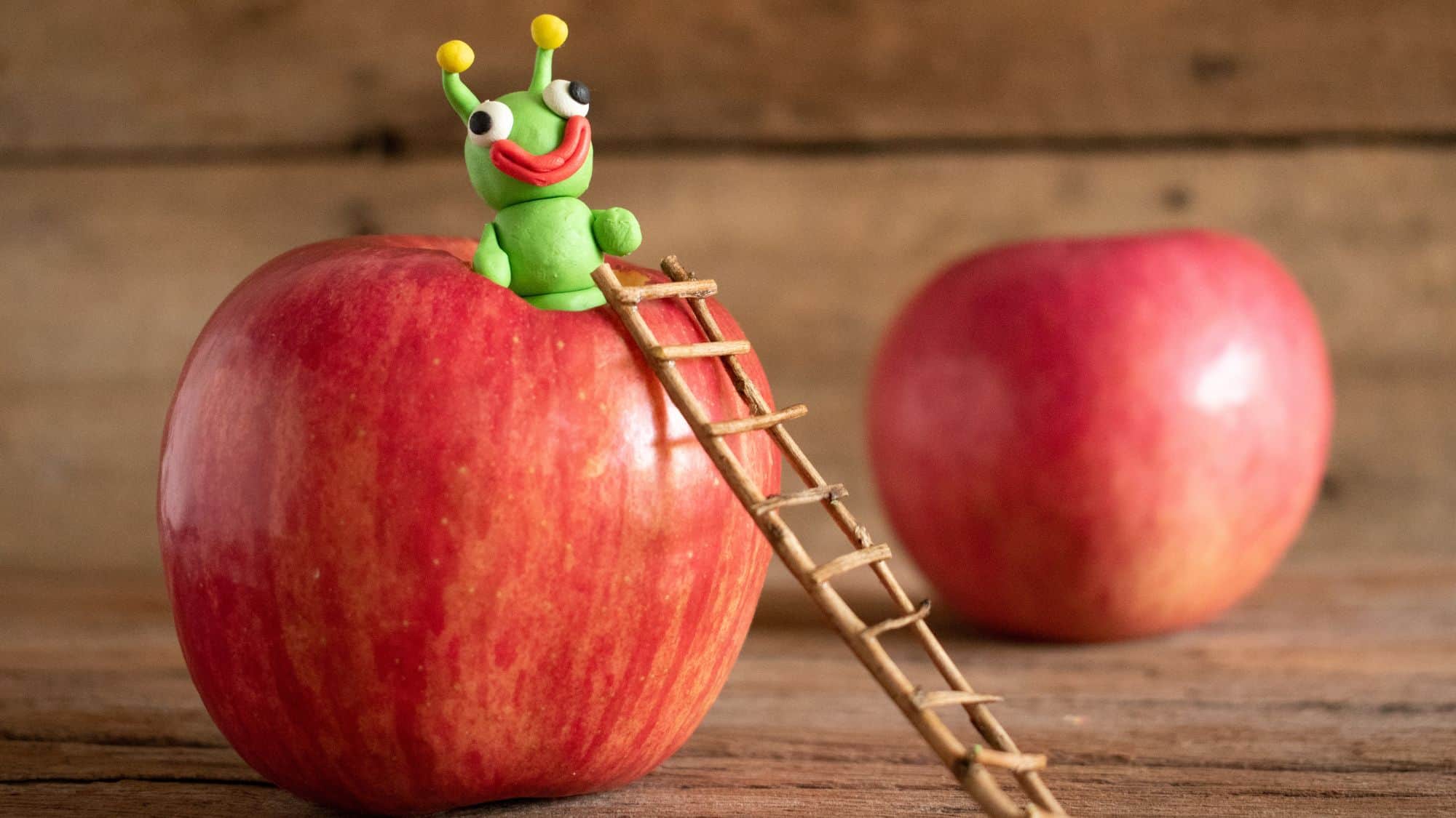 A playful scene featuring a small, green, clay alien figurine with big googly eyes and a wide red smile perched atop a shiny red apple. A tiny wooden ladder leans against the apple, adding a whimsical touch.