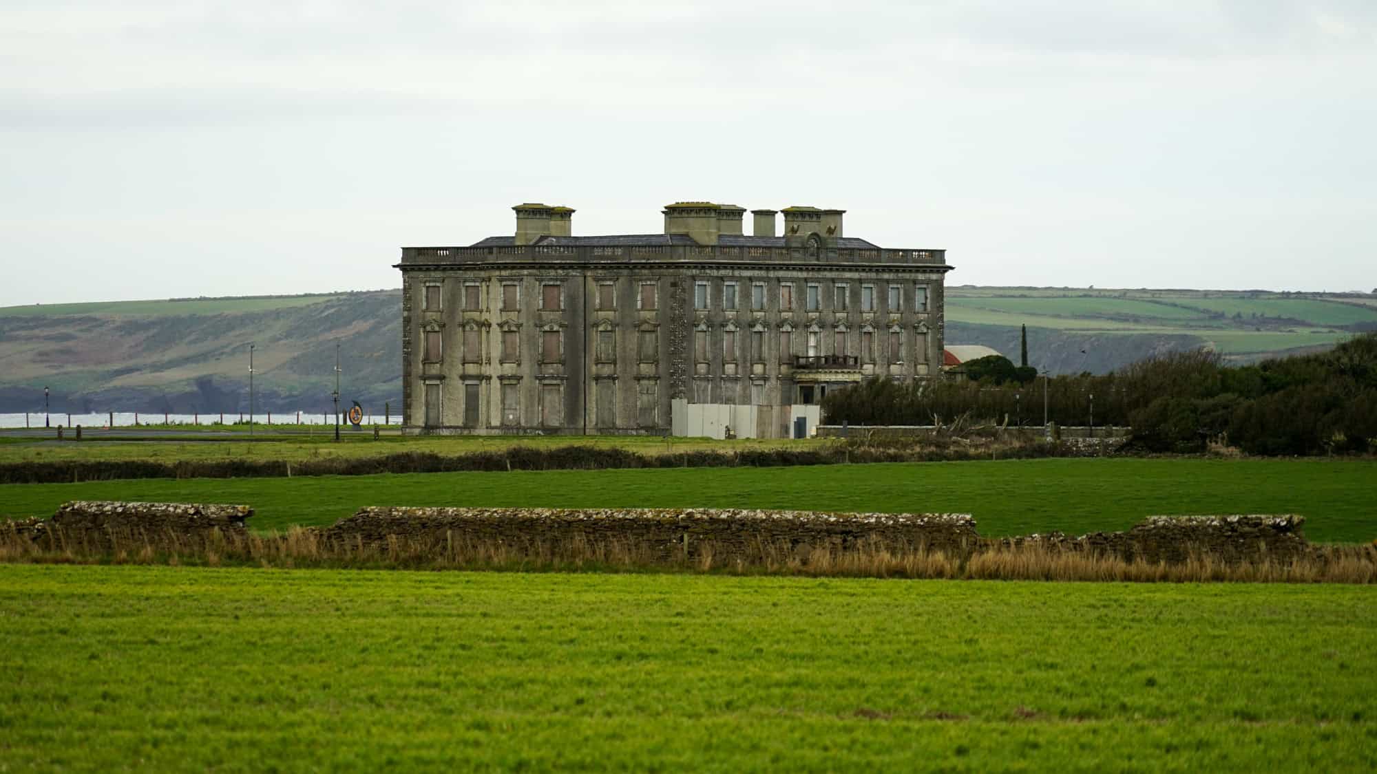 A grand but abandoned Georgian mansion with a gray, decaying facade, standing in an open green field near the coastline. Its many boarded-up windows and neglected structure add to its haunted reputation.